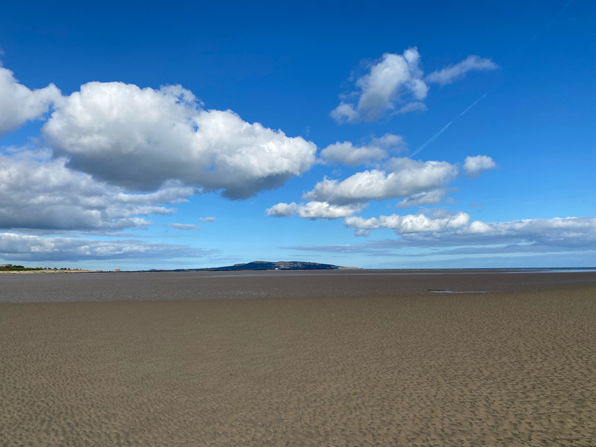 Empty expanse of wet sand on Dublin Bay at a low tide with Howth in the distance and Poolbeg pier and lighthouse visible below a blue sky with a few puffs of white cloud.