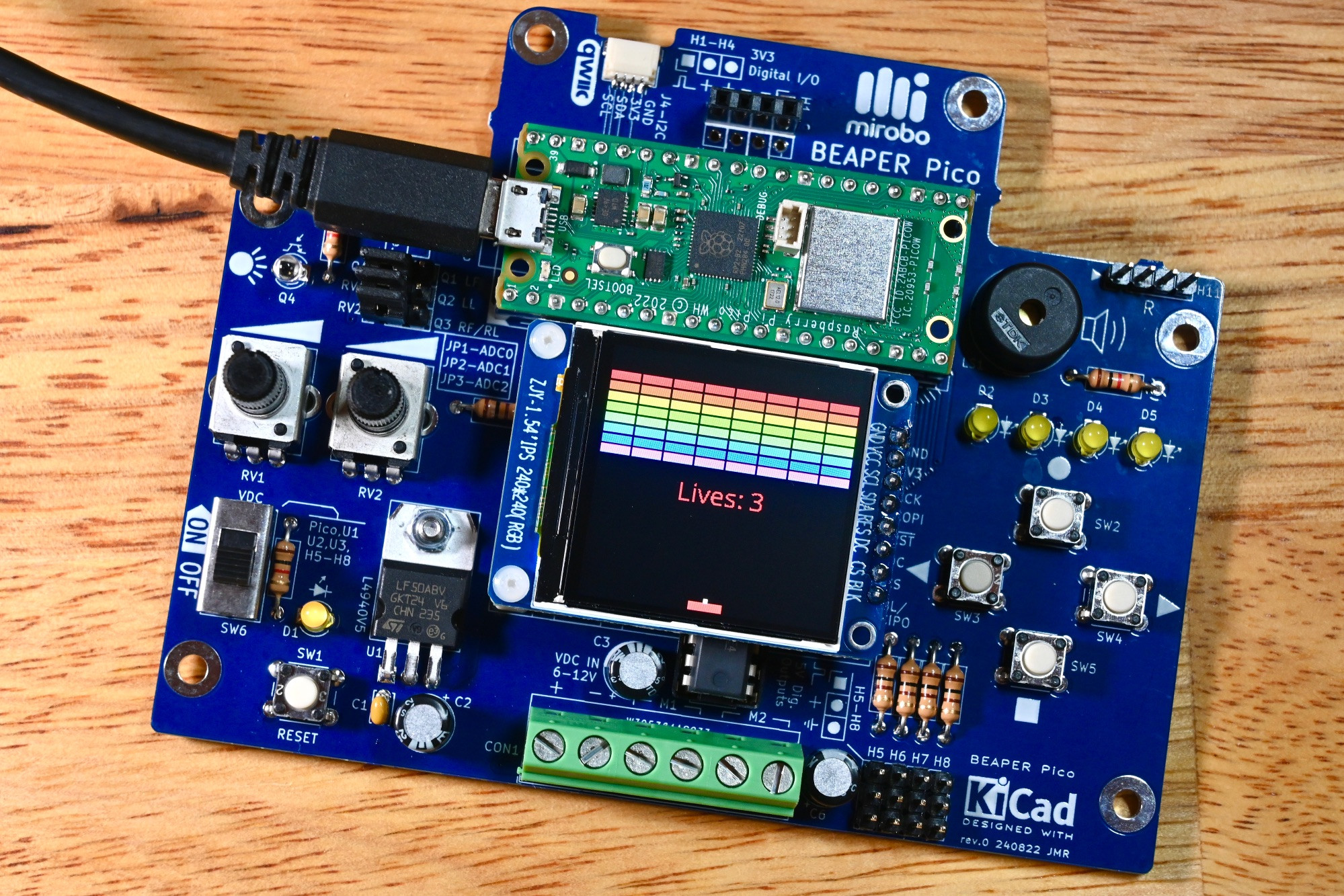 Top view of a fully-populated BEAPER Pico, a beginner education circuit for Raspberry Pi Pico, running a block breaker game on its on-board LCD display. This BEAPER Pico contains (left to right) an ambient light sensor, two user potentiometers, a voltage regulator circuit, a Raspberry Pi Pico W microcontroller, an LCD display, level shifter and motor driver ICs (under the display), a piezo speaker, four user LEDs, four pushbuttons, and four servo connectors. At the very top of the PCB is an I2C QWIIC connector and a 4-pin digital I/O header. The green screw terminal strip at the bottom connects to a battery and motors.