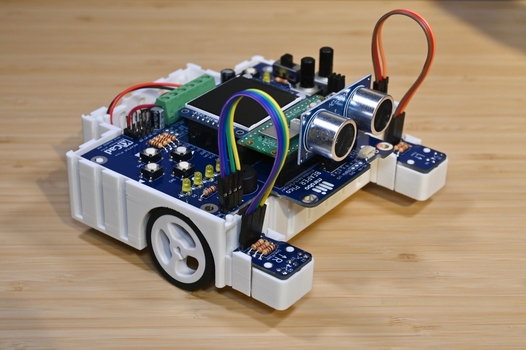 A white 3D-printed BEAPER Bot robot controlled by a BEAPER Pico circuit with an on-board Raspberry Pi Pico W and optional graphical LCD. The robot is built in a front-drive configuration and has two optical floor sensors and a SONAR module installed.