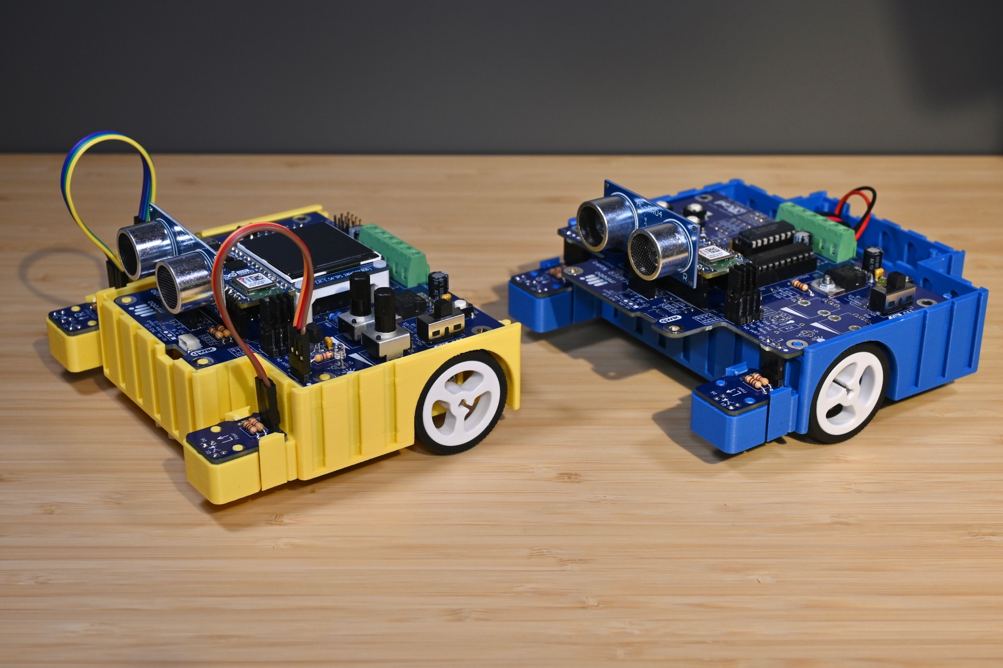 Two 3D-printed BEAPER Bot robots controlled by Arduino Nano ESP32 microcontrollers installed in BEAPER Nano PCBs. The yellow robot (left) is set up in a rear-drive configuration and has an optional LCD installed. The blue robot (right) is set up in a front-drive configuration. Both robots have dual optical floor sensors and a SONAR module installed.