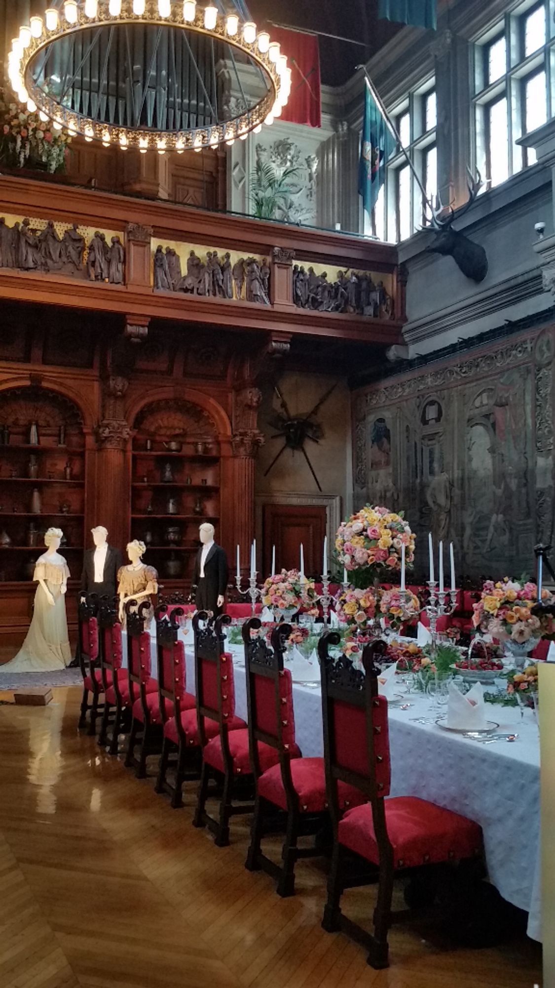 An elegant dining room with minstrel gallery and clerestory windows above. The long table is surrounded by plush upholstered red dining chairs, with white tablecloth, elaborate place settings and flowers in epergnes. Costumed manikins at the head of the table have spotlights on them.