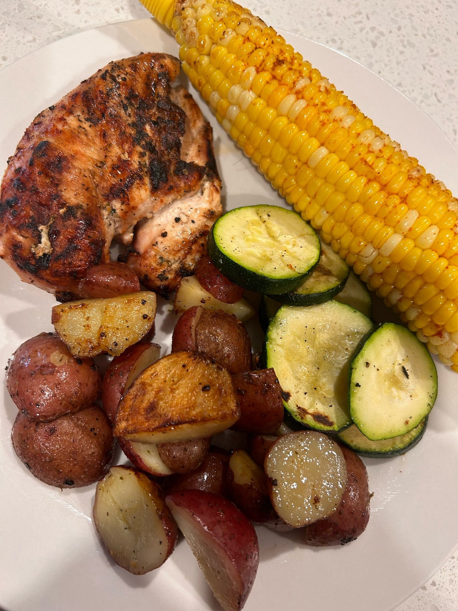 A plate with a bbq chicken breast, potato, zucchini and corn