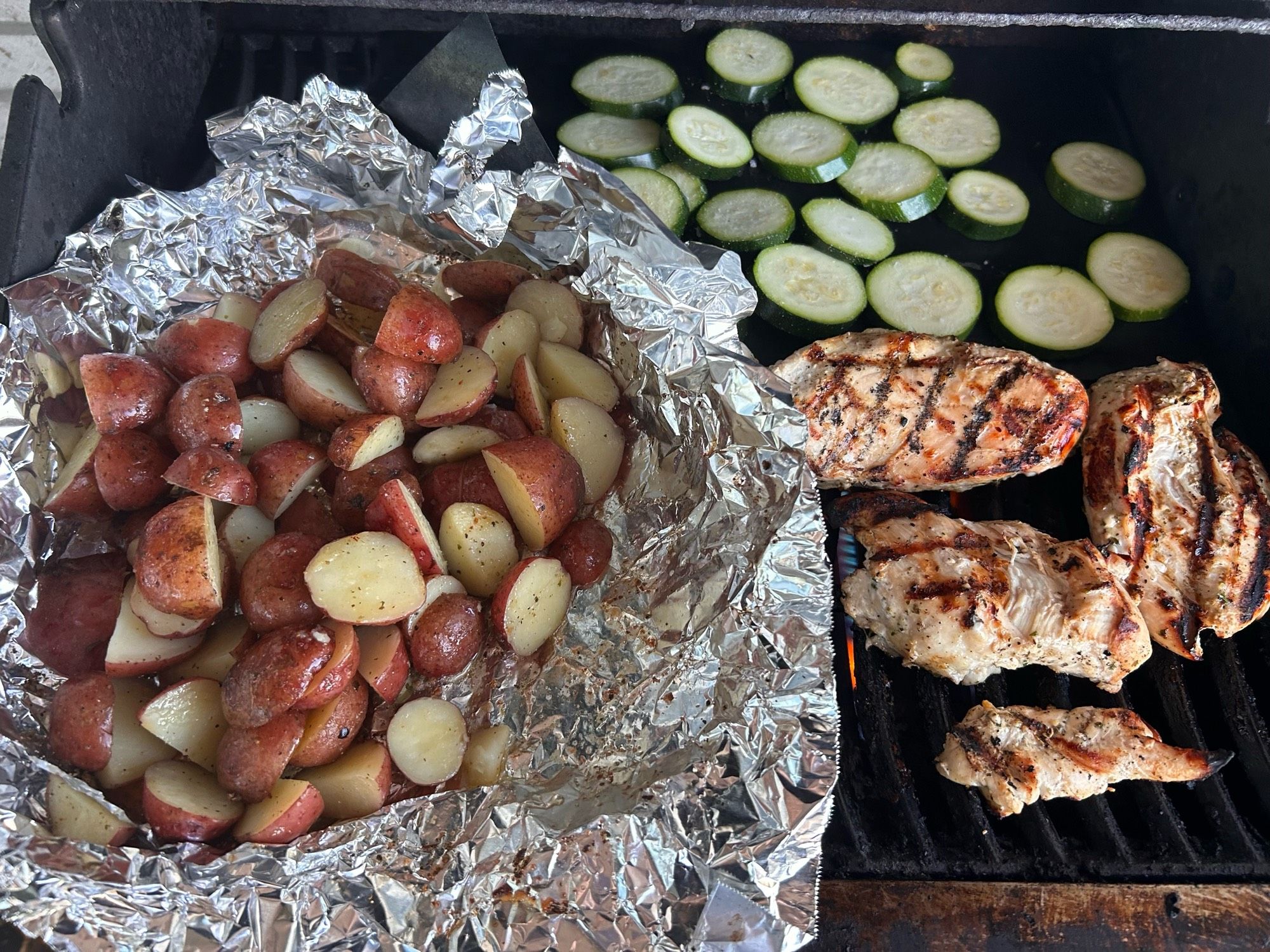 A bbq with a foil pack of potatoes, on one side, chicken breast on the other and sliced zucchini in the back