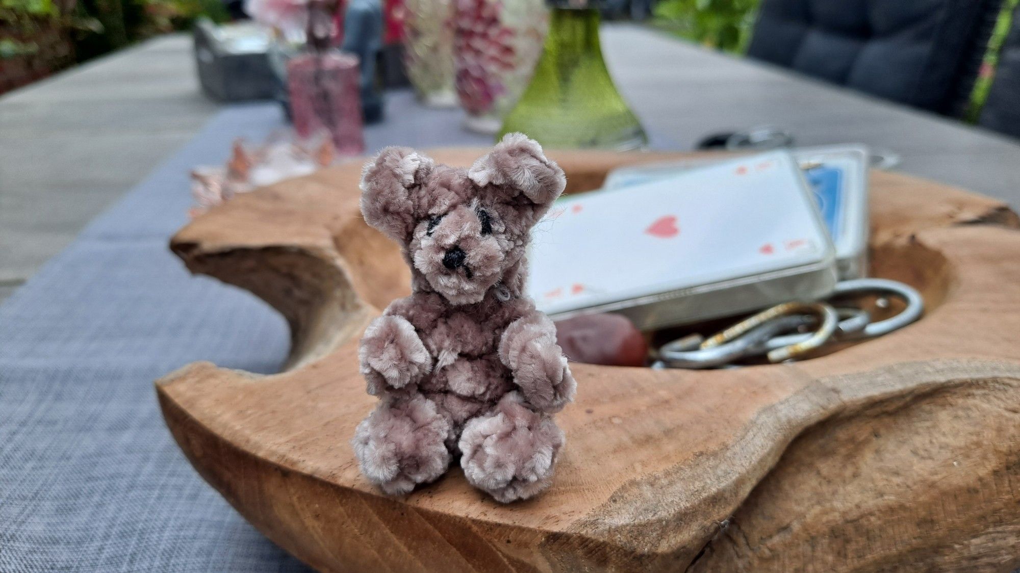 A very small light brown teddy bear crocheted with fuzzy yarn. It's sitting on the rim of a wooden bowl on an outdoor table.