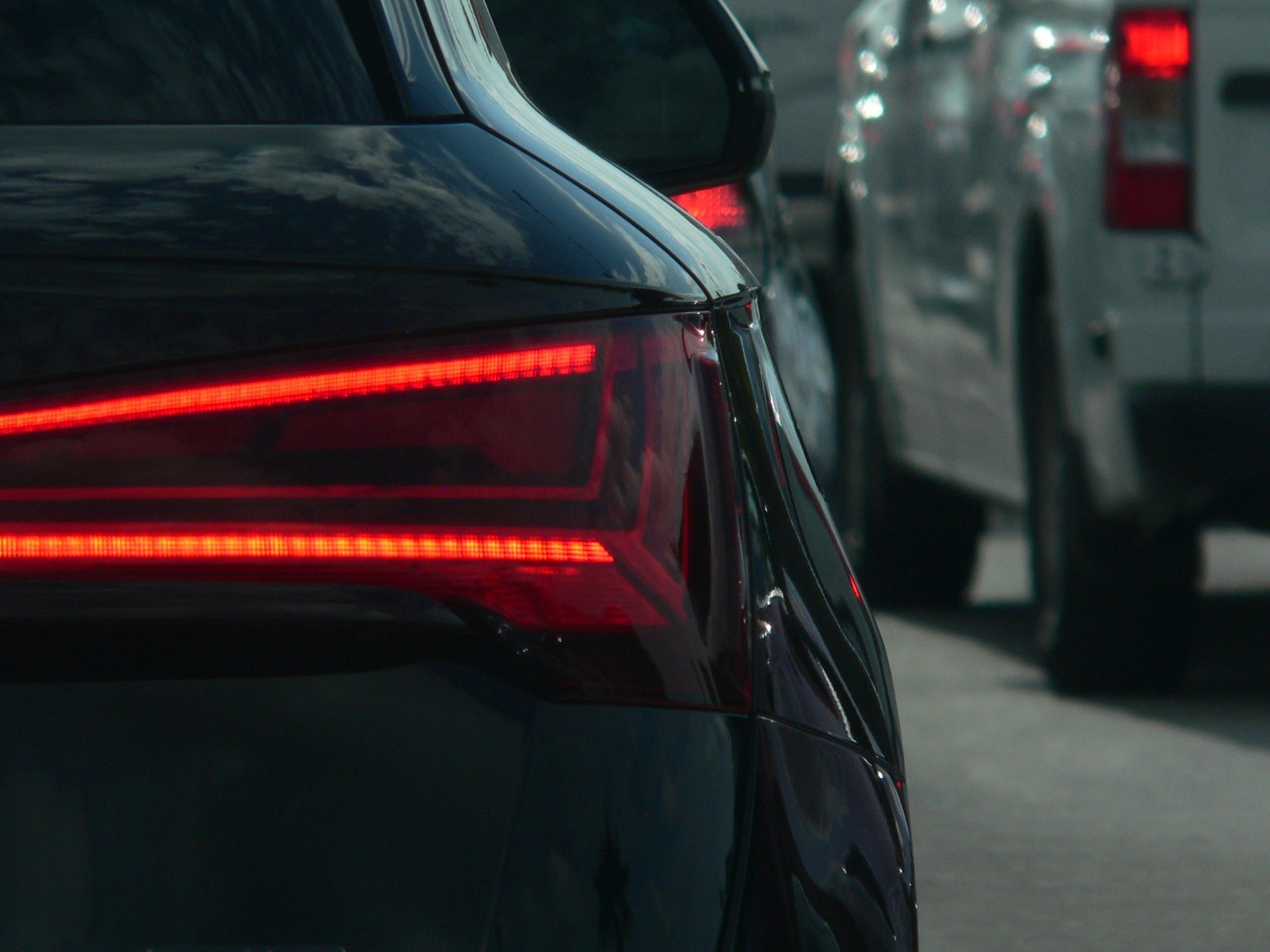 Left 2/3 of frame are a close shot of a dark grey Audi SUV's led taillight lit up, and the right 1/3 of frame is a white f-150 pickup out of focus