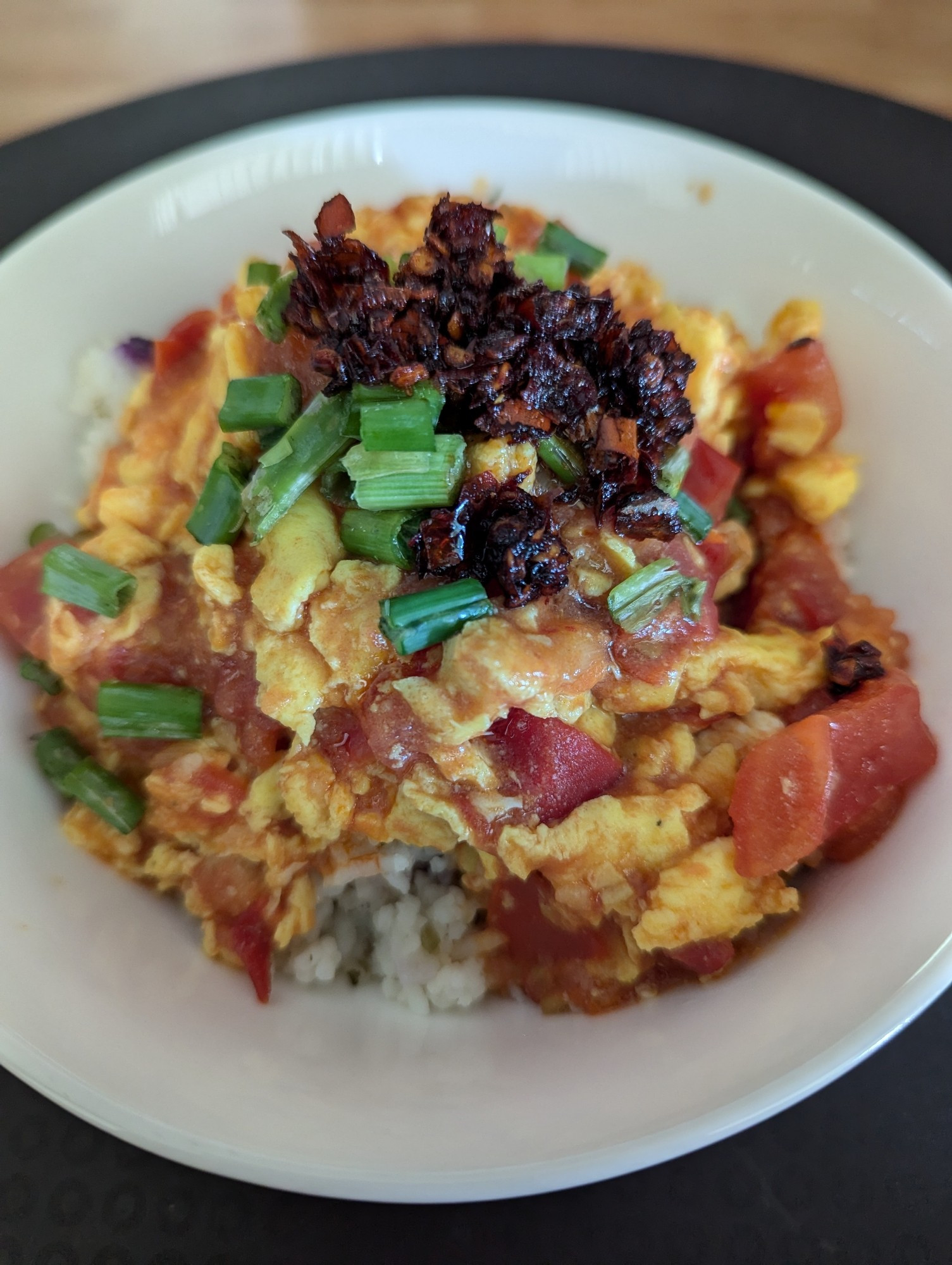 Tomato and eggs on a bed of rice with green onions and chili crisp sprinkled on top all sitting in a white bowl