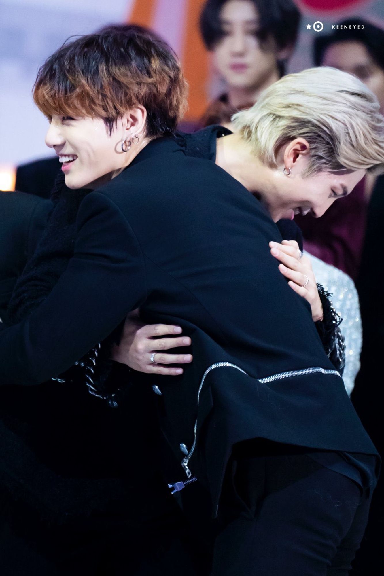 Jungkook and Namjoon hugging at an awards show. Jungkook has ombre brown hair and Namjoon has blond. Namjoon is only seen in profile over Jungkook's shoulder, bent over, and both are smiling broadly.