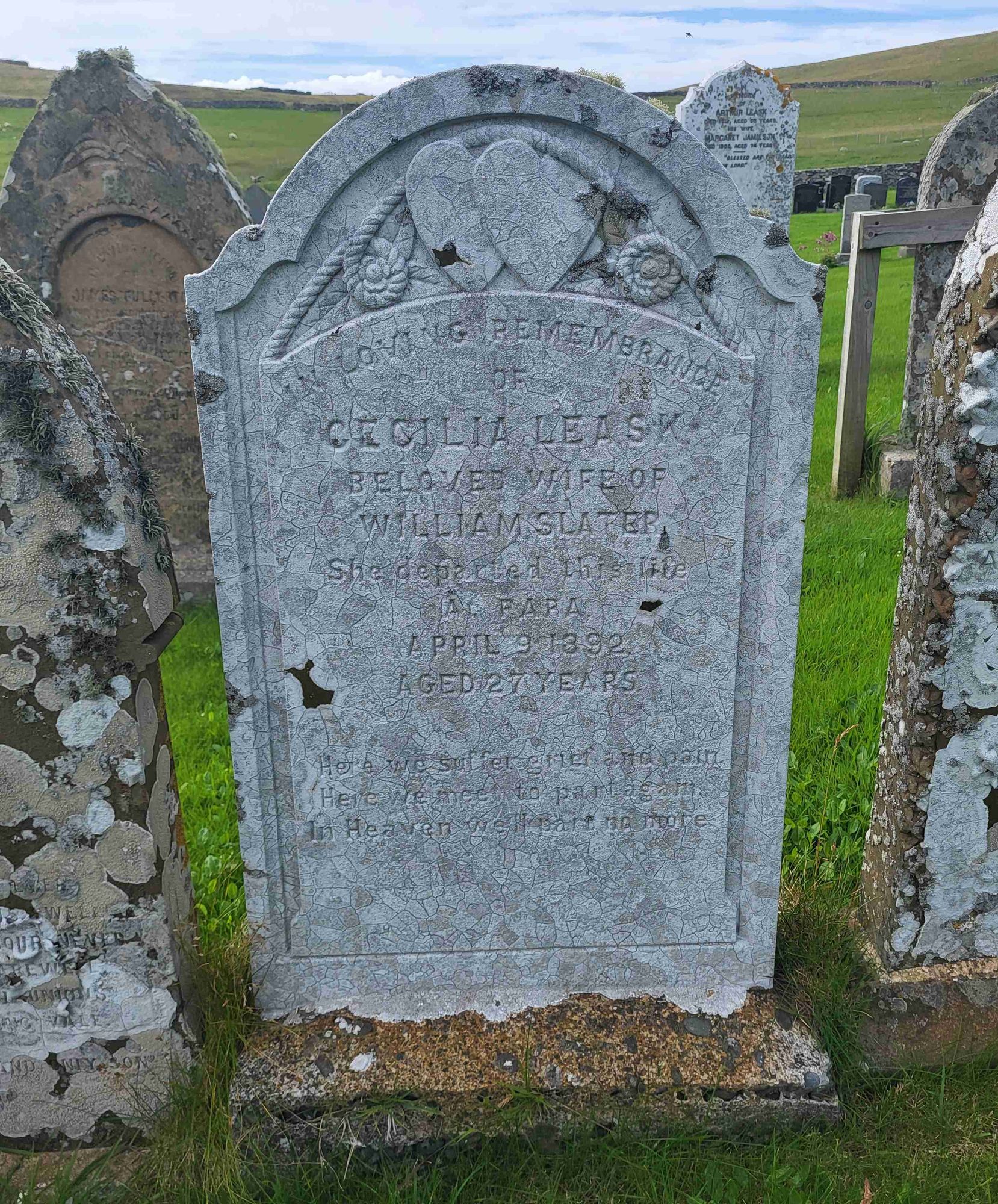 A sandstone gravestone covered in hard white lichen. The design at the top shows 2 hearts with a coiled rope around them. The stone is dedicated to Cecilia Leask, 1892.