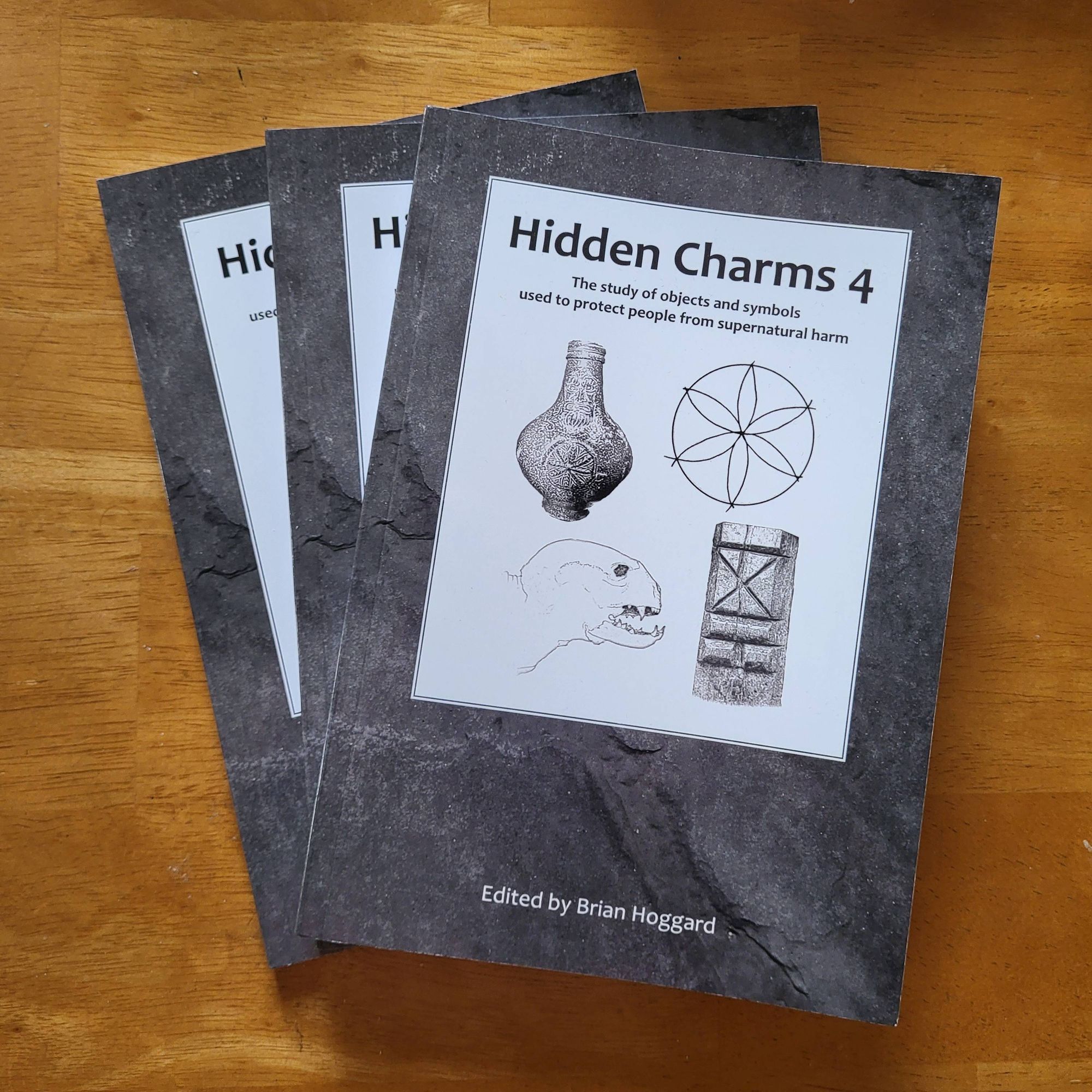 Three grey books, titled 'Hidden Charms 4', with four images in a white box on the cover. They are a bellarmine jug, a hexfoil, a drawing of a dried cat, and a witch post.