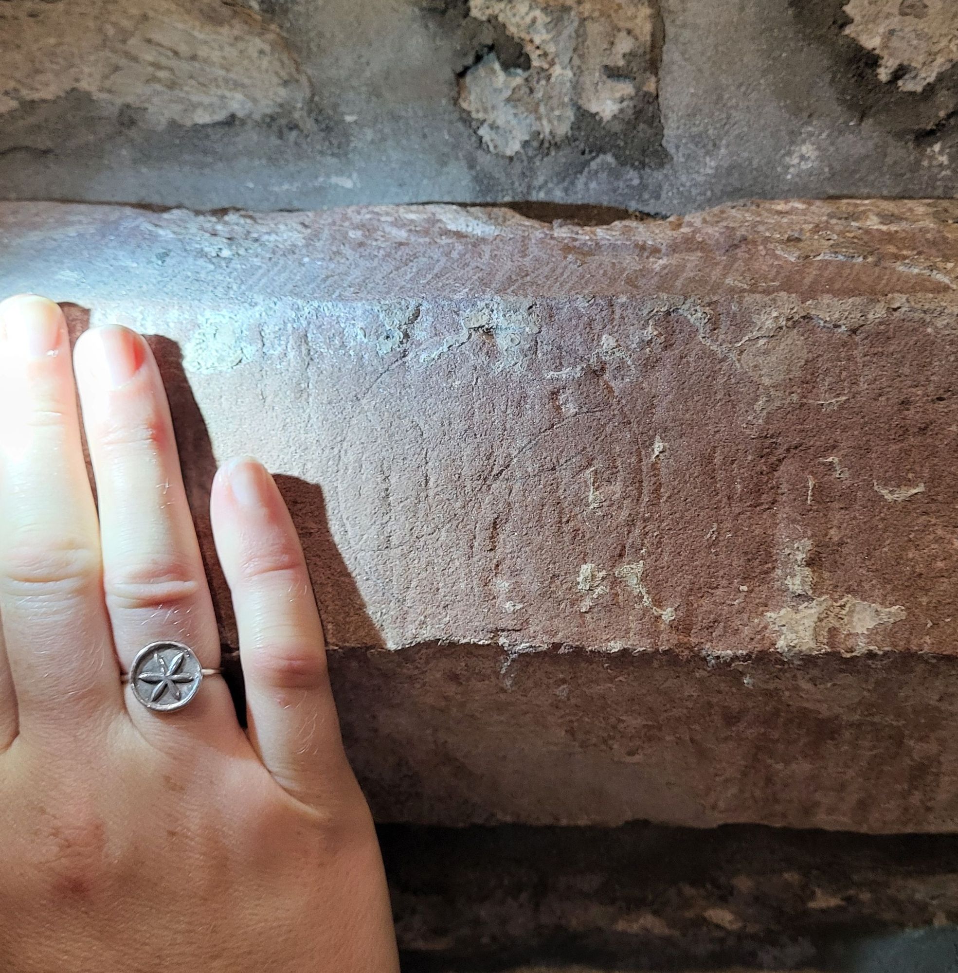 Robyn's hand is resting on the wall, with a silver ring showing a six-petal rosette or hexfoil. On the red sandstone wall is a hexfoil carved in lightly.