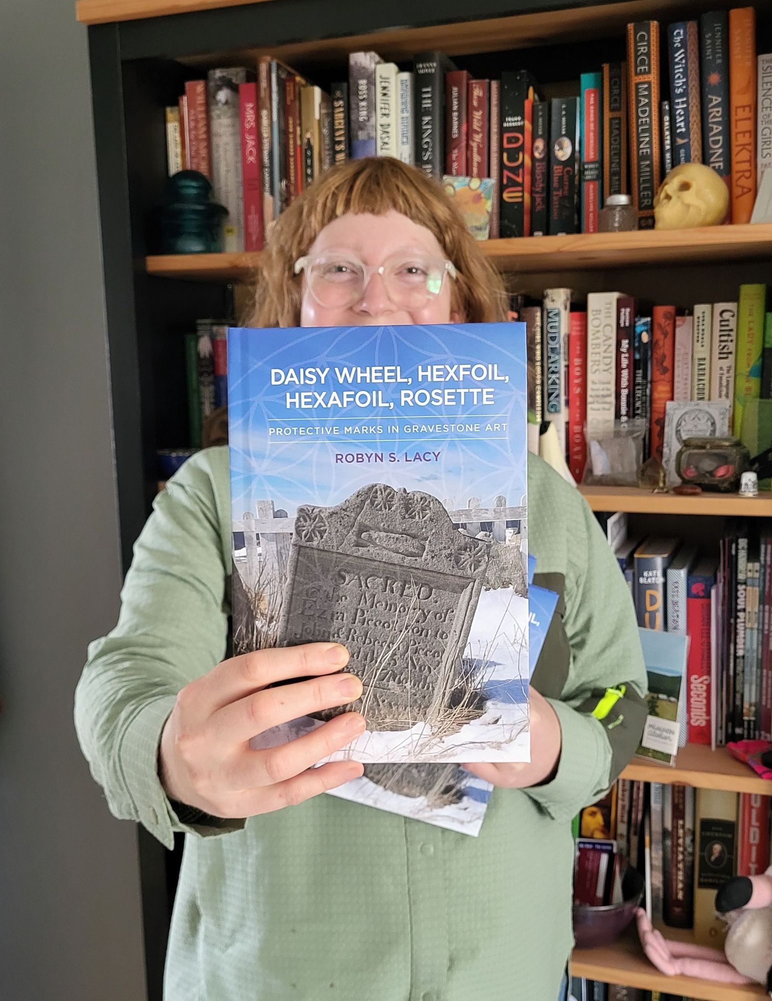 Robyn holds out a copy of her new book, "Daisy Wheel, Hexfoil, Hexafoil, Rosette: Protective Marks in Gravestone Art". She has ginger hair with short bangs and in wearing a green shirt.