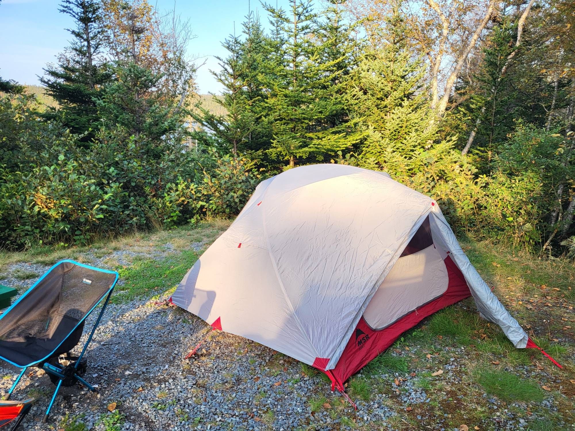 A cute, silver and red MSR tent in a campsite, with a blue and black camp chair on the left. Behind the tent are fir trees and a glimpse of a lake in the background under a blue sky.