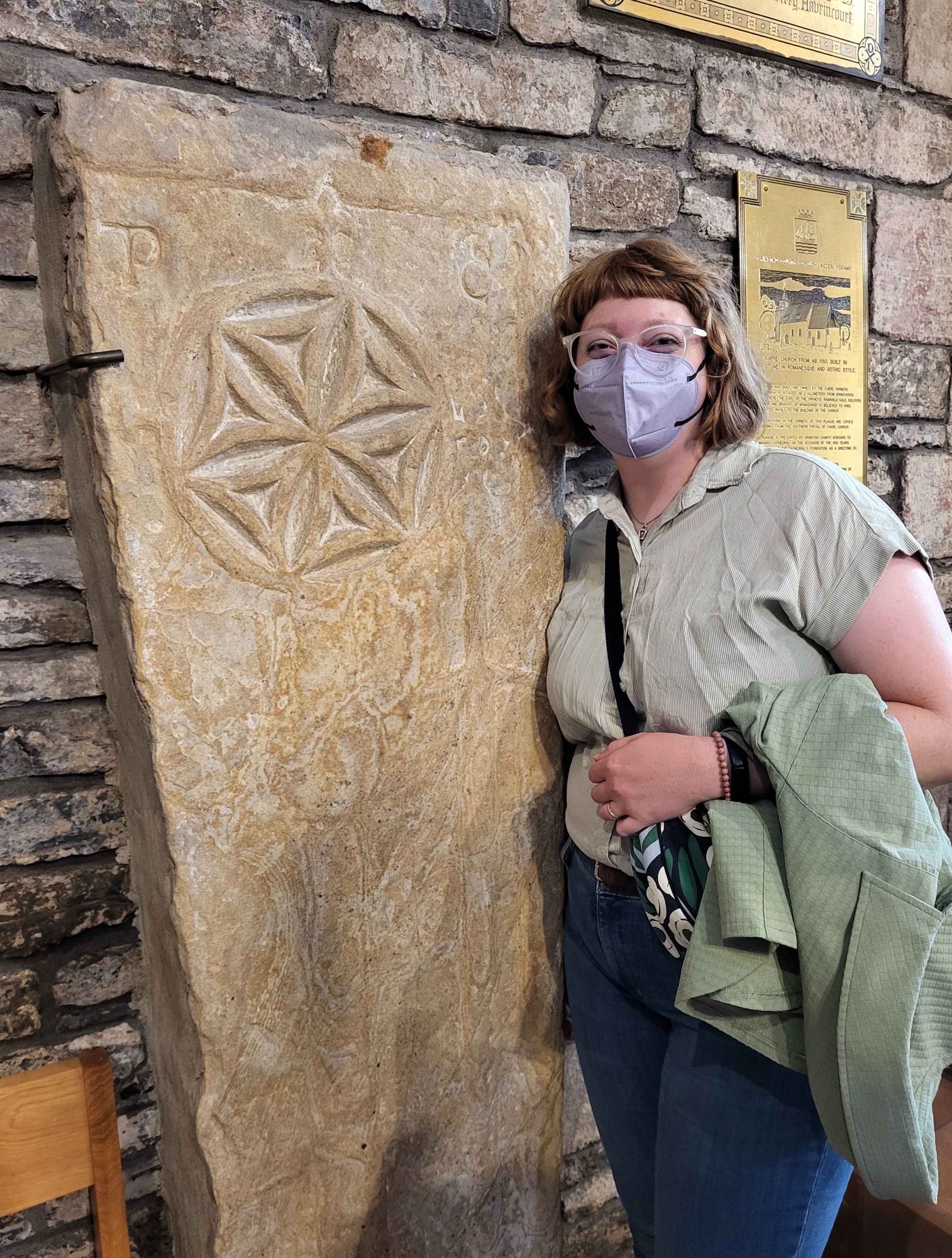 Robyn is standing against the wall, beside a tall grave slab from the 13th century, that has a six-petal rosette or hexfoil at the top of a stepped rod. There is a sword carved beside it. Robyn has red hair with short bangs and bobbed hair, and is wearing white glasses and a purple face mask, and a green button up shirt and jeans.