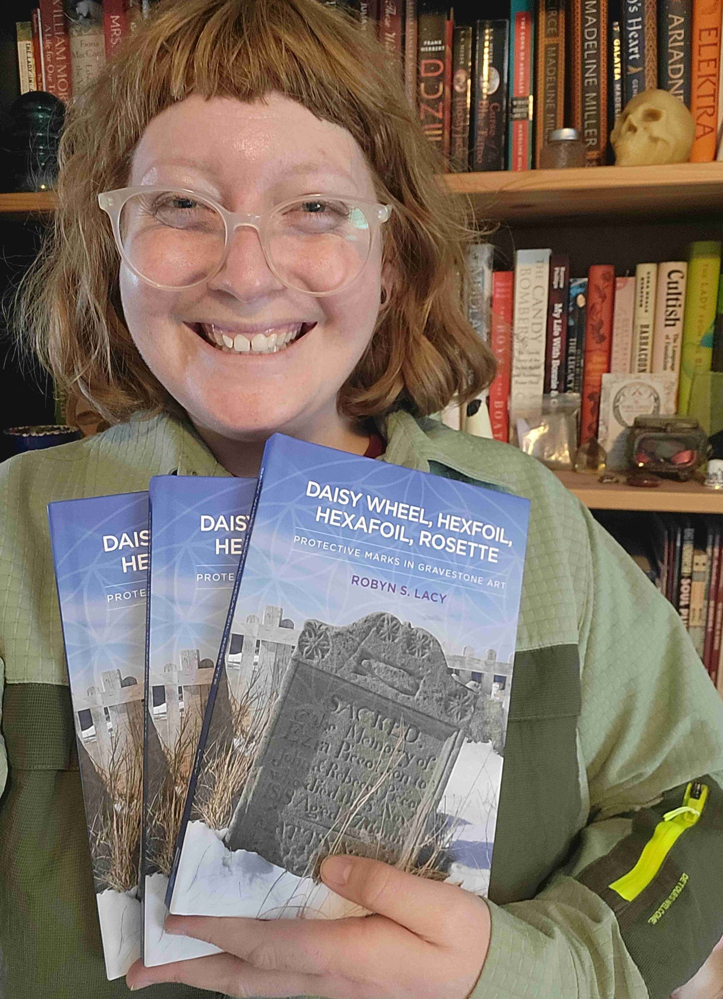 Robyn holds 3 copies of her new book, "Daisy Wheel, Hexfoil, Hexafoil, Rosette: Protective Marks in Gravestone Art". She has ginger hair with short bangs and in wearing a green shirt.