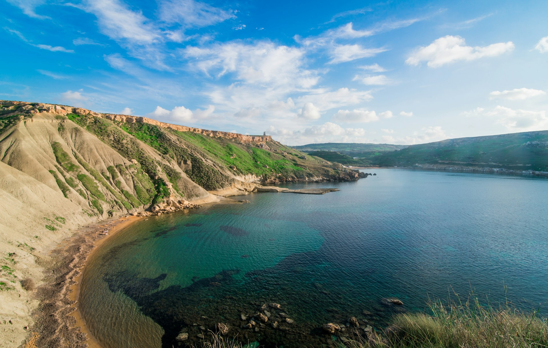 A rugged coastline with green hills, rocky formations, and clear blue waters typical of Malta's scenic coastal landscape.