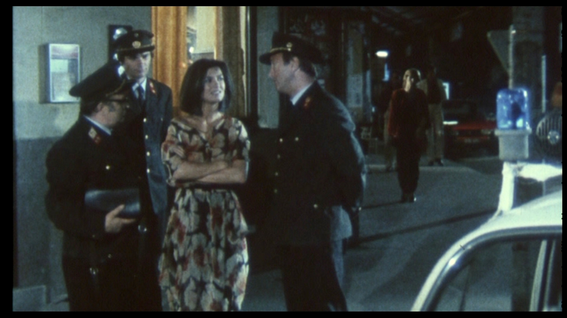 Filmstill aus Karambolage von Kitty Kino: eine junge Frau im Kleid, umringt von drei Uniformierten vor dem Kaffeehaus des Verbrechens. Einer hält ihre Tasche in der Hand. Es ist finster. Im Hintergrund die Freund*innen der Hauptfigur.