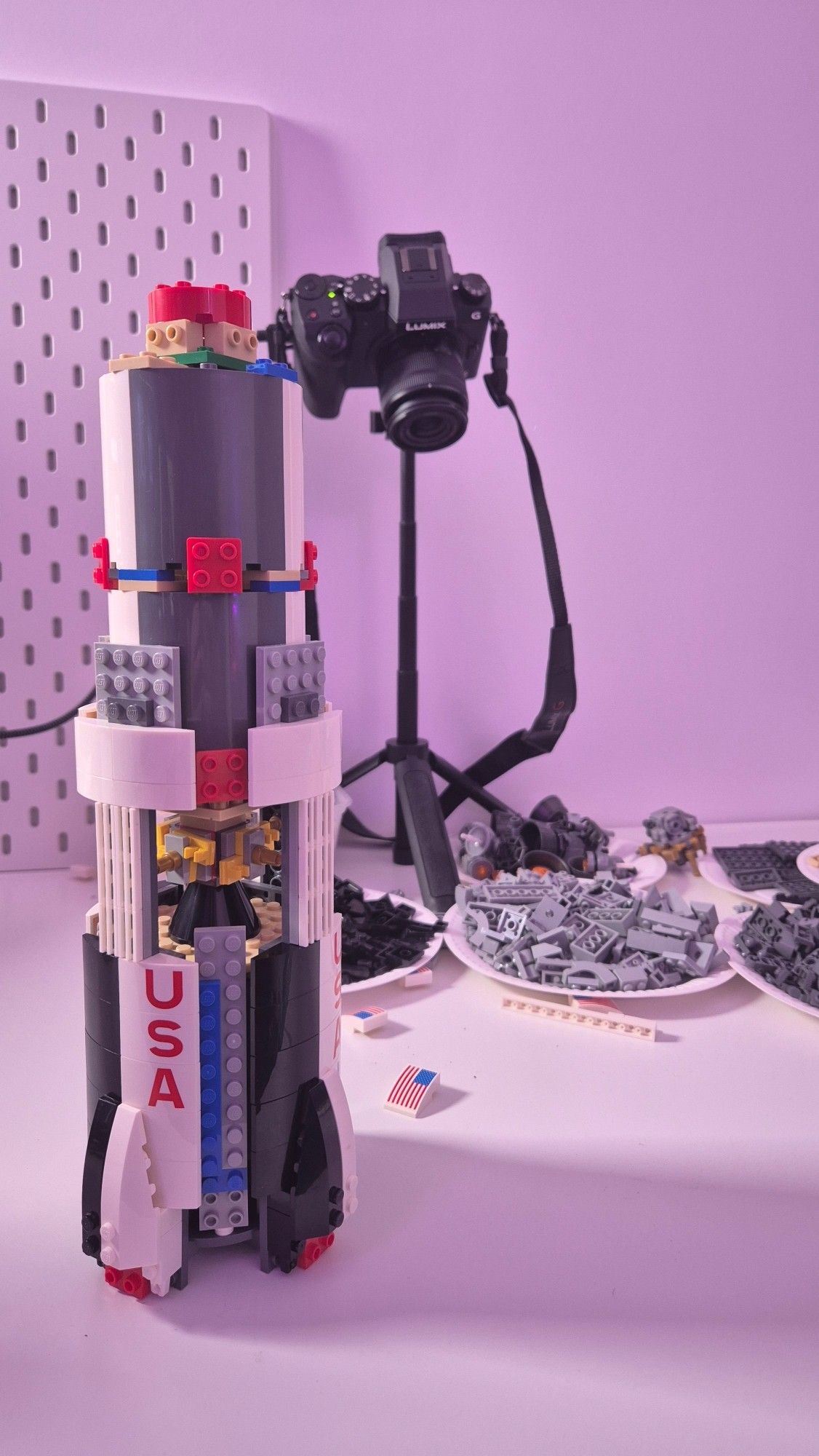 Une Saturn 5 en lego à moitié montée en premier plan, et en second plan une caméra qui filme le montage