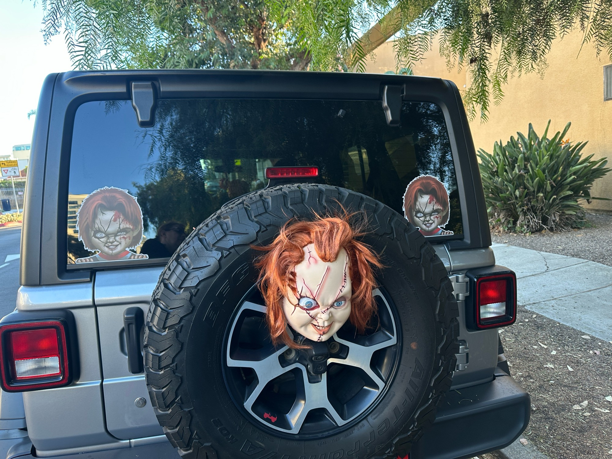 Rear view of a Jeep with Chucky face stickers on both sides of the rear window and a big full color 3D Chucky mask adorning the spare tire, so it would stare menacingly at you as you drive behind it.