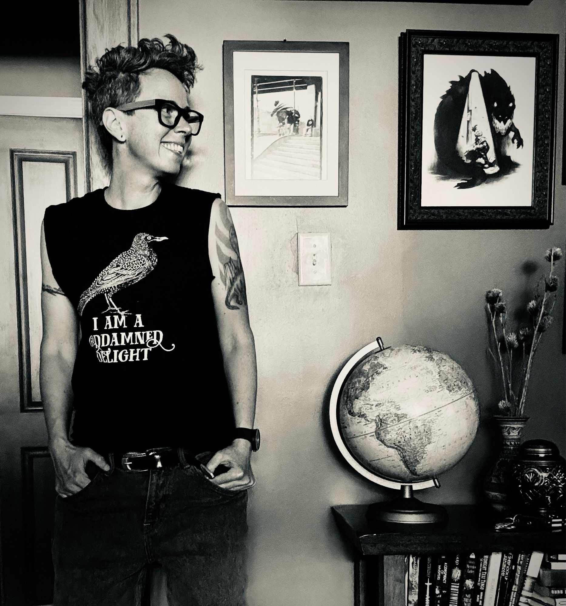 black & white photo

a white trans man stands in a doorframe smiling off to the right. he is in the right of the frame, hands in pockets of grey jeans, a black muscle tee on with a starling (Effin Birds) that says “I Am A Goddamned Delight”

the background is full of black and white framed art above a globe, dried thistles, and an urn on a low bookshelf