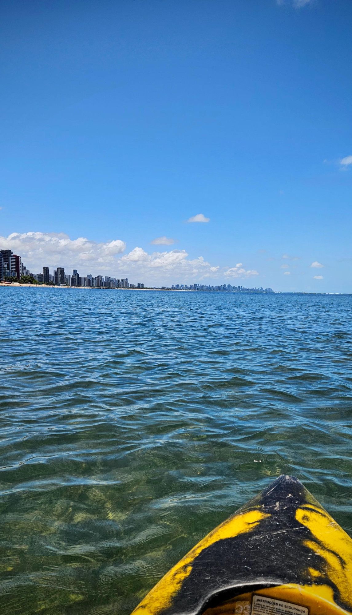 Foto contendo a ponta de um caiaque em um mar azul meio cristalino, céu praticamente sem nuvens ao fundo com a praia e alguns prédios no horizonte.