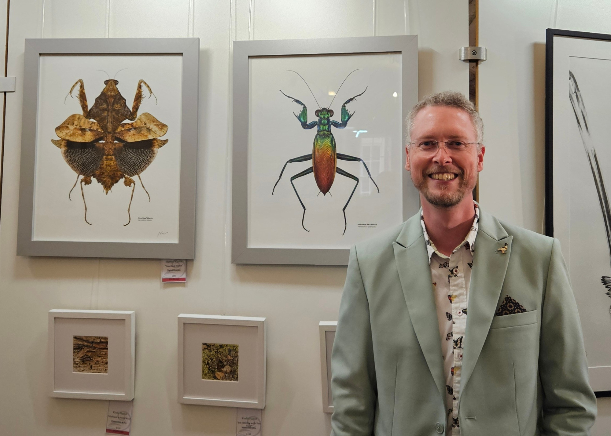 A Really Very Proud Indeed Martyn standing in front of 2 mantis drawings at the Southern Nature Art Exhibition. The mantises shown are the brown dead leaf mantis and the rainbow coloured iridescent bark mantis.