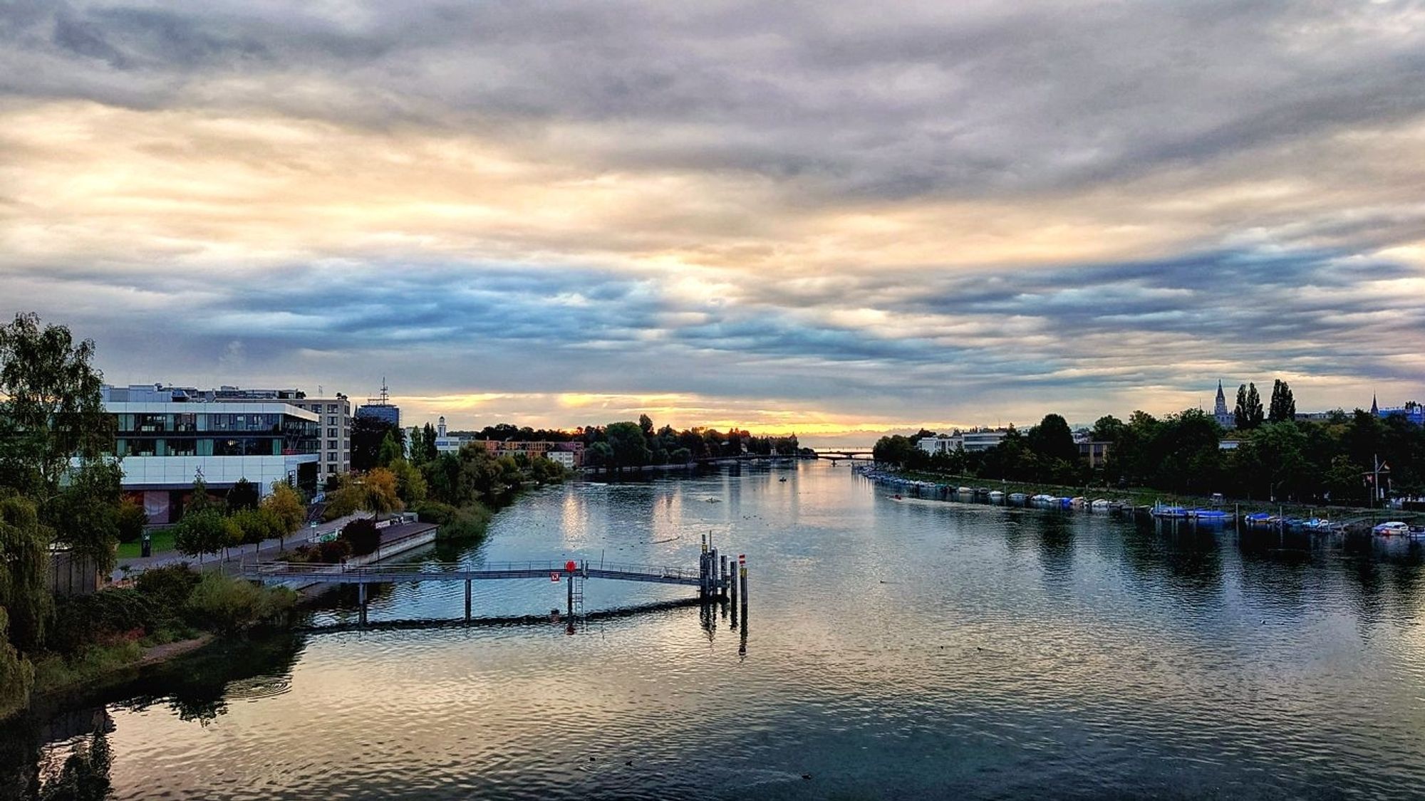 Rheinkilometer 1 in Konstanz. In der Mitte ist der Rhein. Darin steht eine Schiffsanlegestelle, links davon am Ufer ein Mehrzweckgebäude. Rechts sind Boote am Ufer festgemacht. An beiden Seiten des Ufer stehen Gebäude. Am Horizont ist der Bodensee zu sehen sowie zwei weitere Brücken.