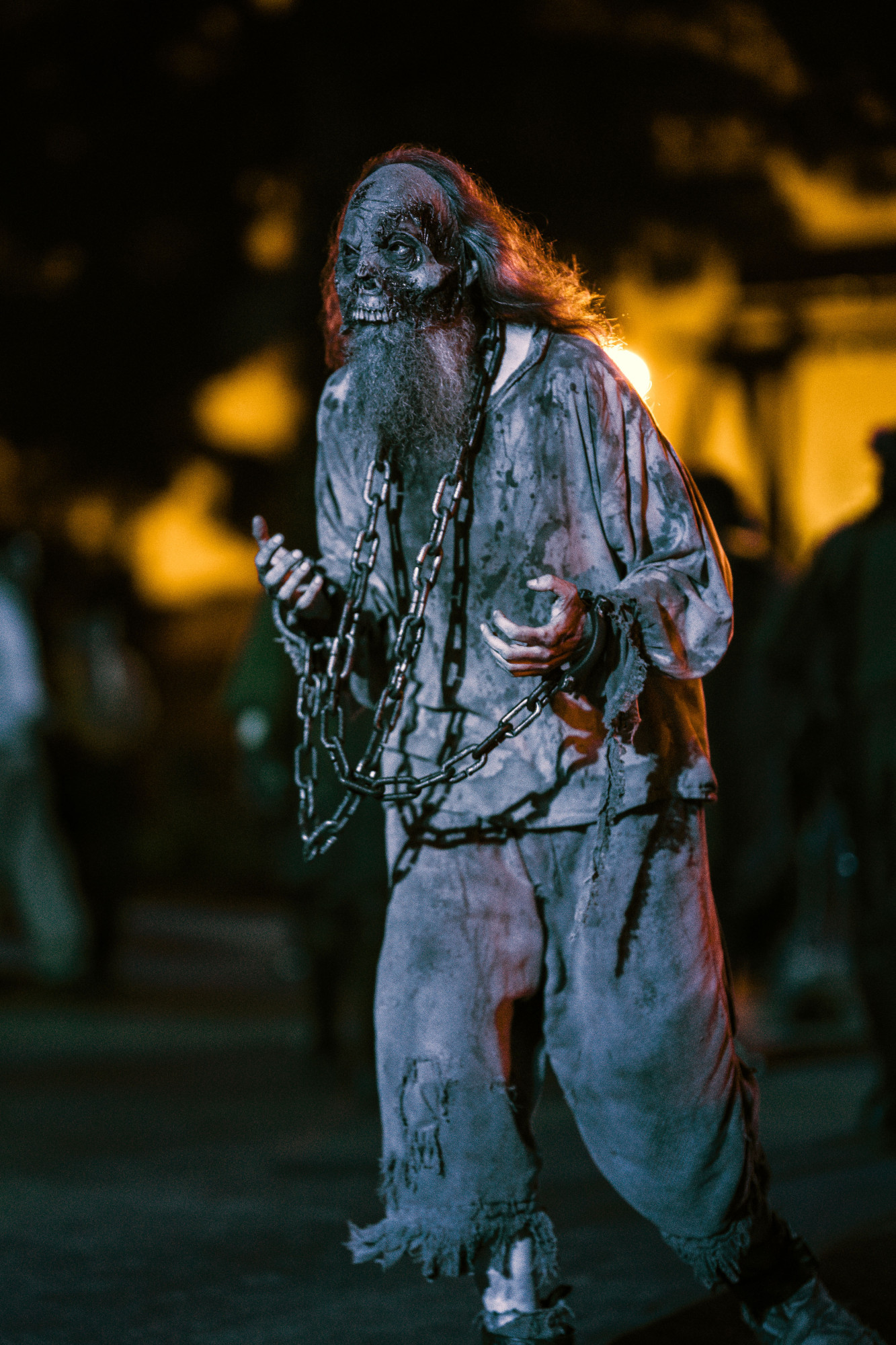 Nighttime photograph by Chris Cornish showing an old tortured prisoner with scraggly long hair and beard dressed in bloodied rags, wearing shackles and draped in chains staggering through a medieval-themed ScareZone in the Knott's Scary Farm attraction.