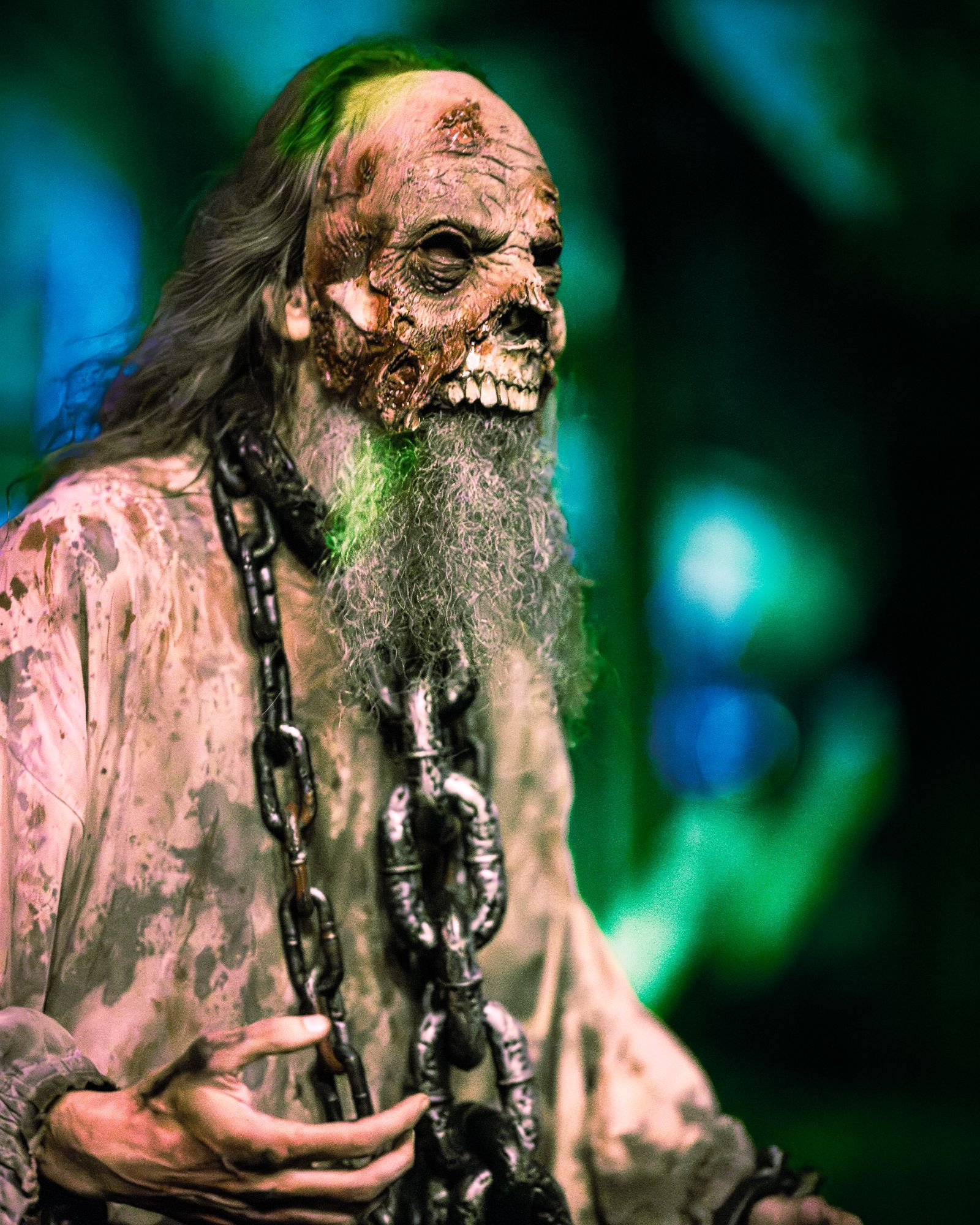 Nighttime photograph by Chris Cornish showing the upper body of an old tortured prisoner with scraggly long hair and beard dressed in bloodied rags, wearing an iron collar and shackles and draped in chains staggering through a medieval-themed ScareZone in the Knott's Scary Farm attraction.