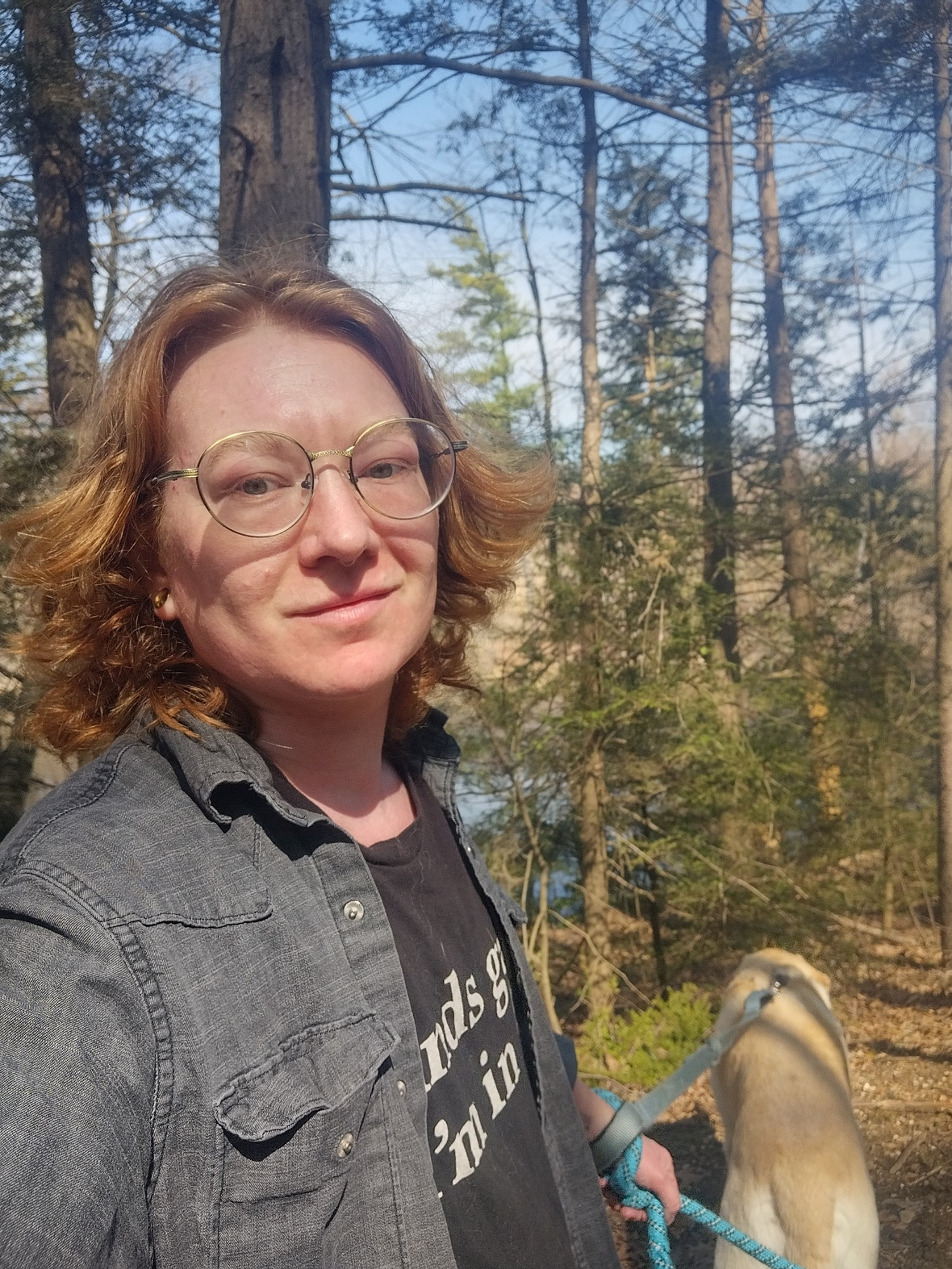 A selfie showing me (V) outside on a woodland walk holding two blue leashes. One dog is visible in the background, a white lab mutt. I have shaggy red hair and am wearing glasses, a black denim overshirt, and a black tee that says "sounds gay, I'm in." 