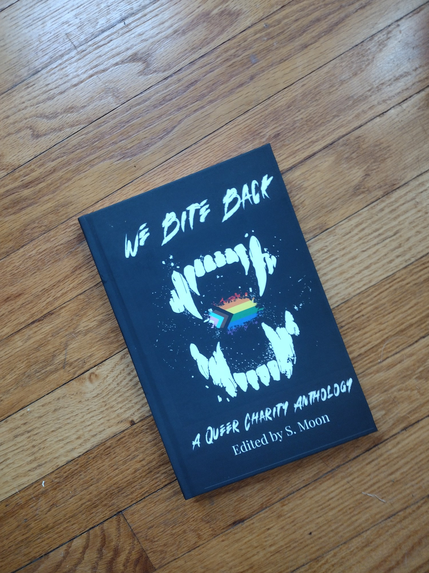 A hardcover copy of WE BITE BACK a queer charity anthology on a hardwood floor. The cover art is a fanged mouth showing the progress pride flag inside. It is edited by S. Moon. 