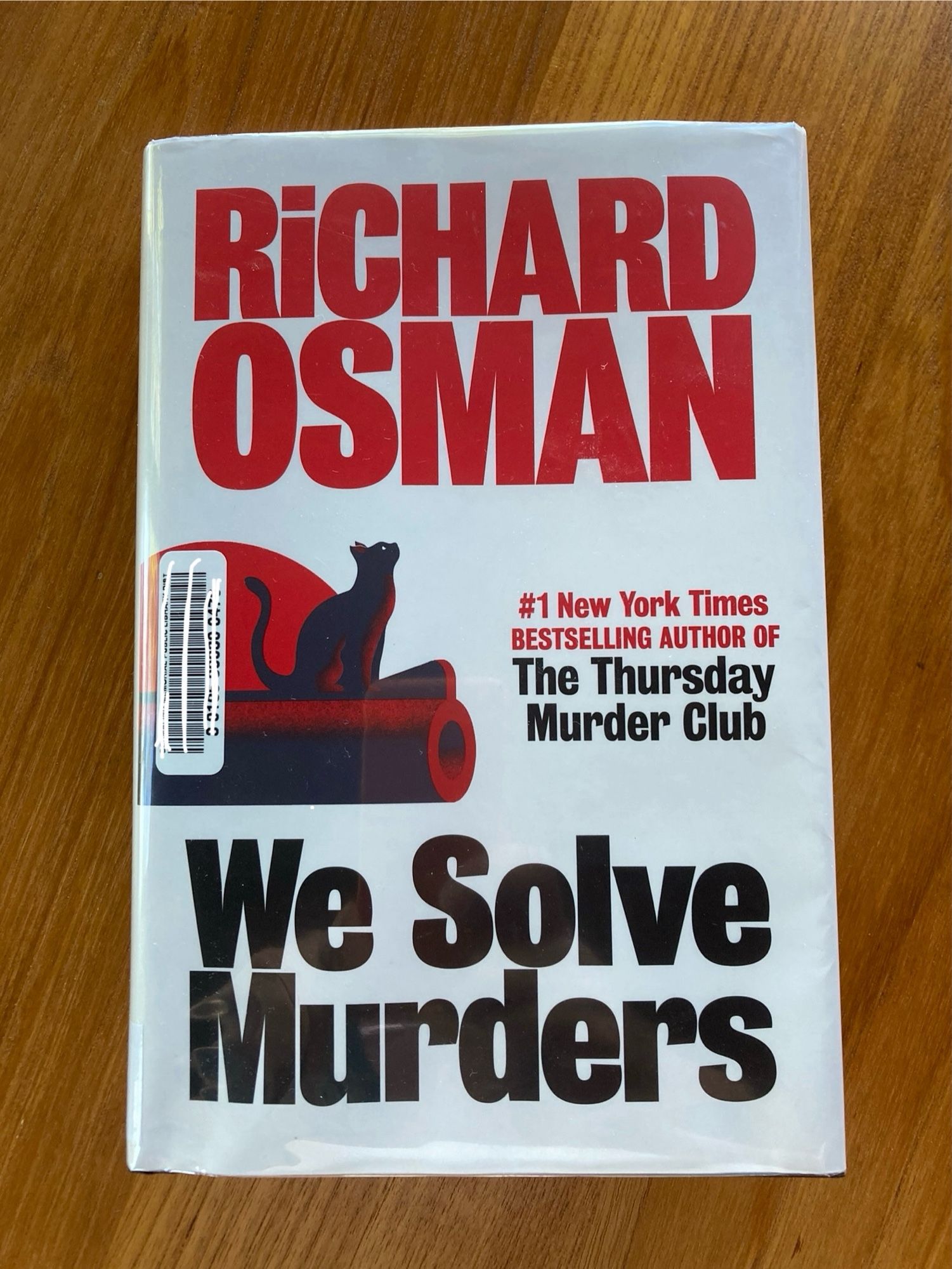 Cover of the book WE SOLVE MURDERS by Richard Osman. It has a white background. The author’s name is in red and the title is in black. There is a black cat on the barrel of a red gun.