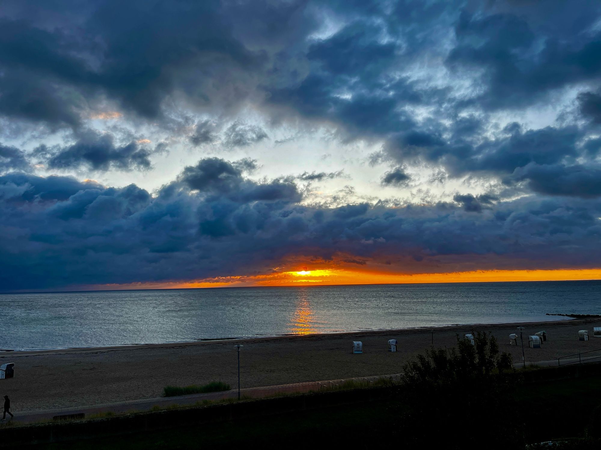 Blick Richtung Sonnenaufgang. Es spiegelt sich nur leicht die Sonne und sie ist zu erahnen durch einen leichten roten Schimmer. Ansonsten viele Wolken. Im Vordergrund der gewohnte Strandabschnitt.