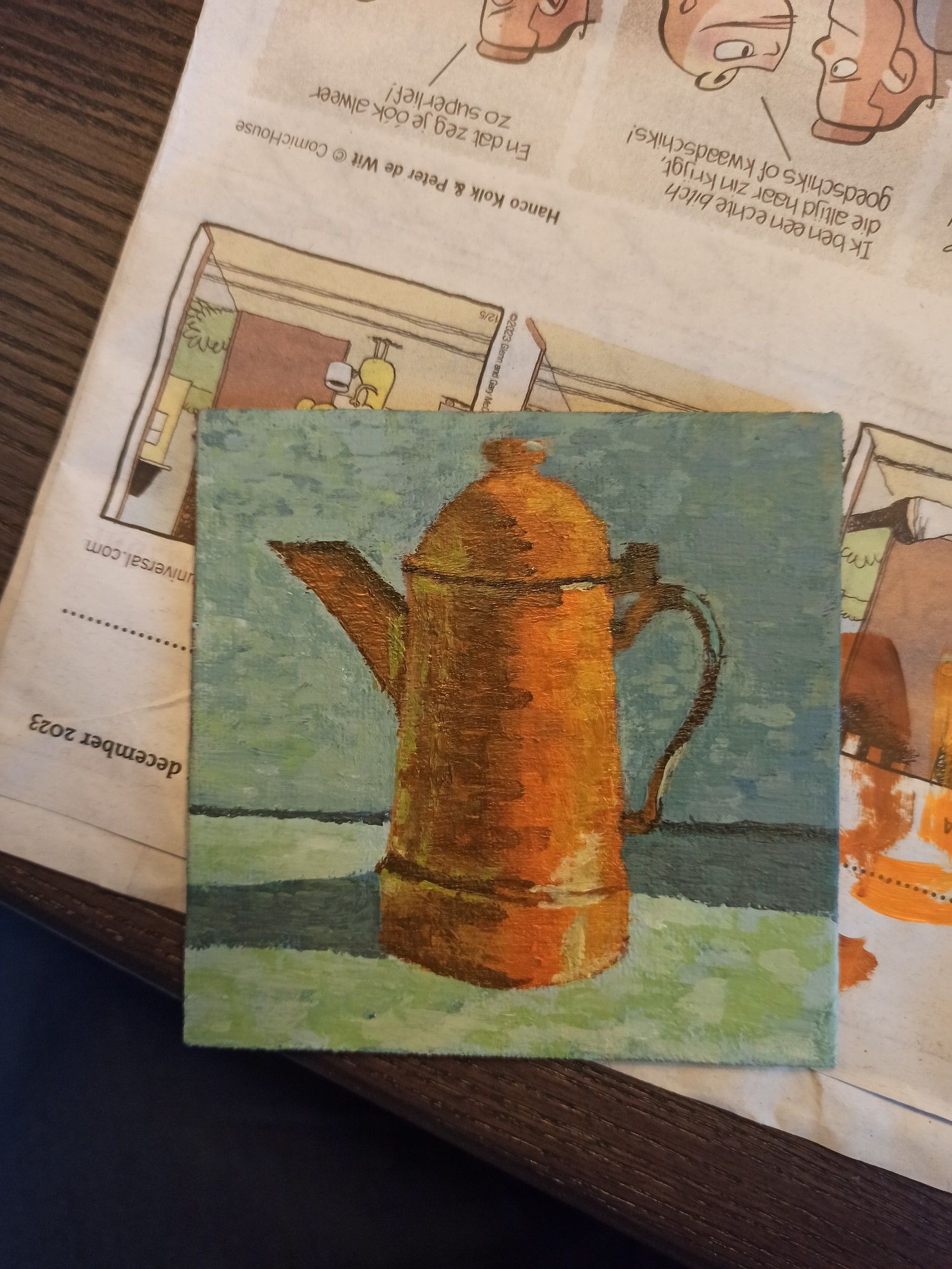 On top of newspaper, lays a small acrylic painting of a tall orange kettle with grey background