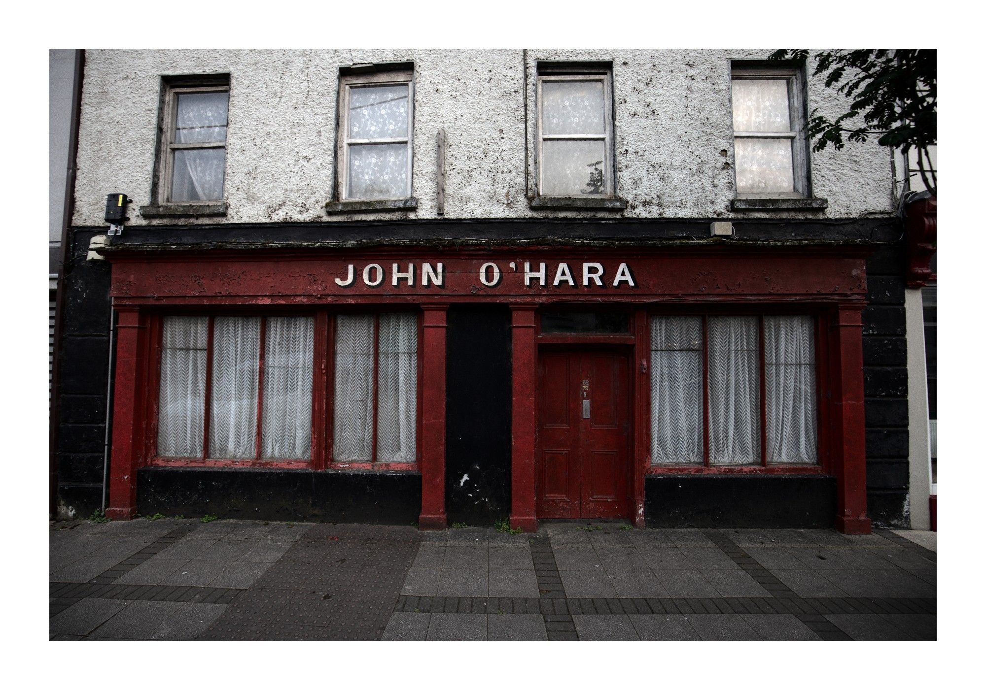 An old, weathered building with a faded red facade features the name "John O'Hara" in white letters above the door. The windows are covered with lace curtains, and the exterior shows signs of age, with peeling paint and worn walls. The building has a quaint, rustic feel, evoking a sense of history and nostalgia.