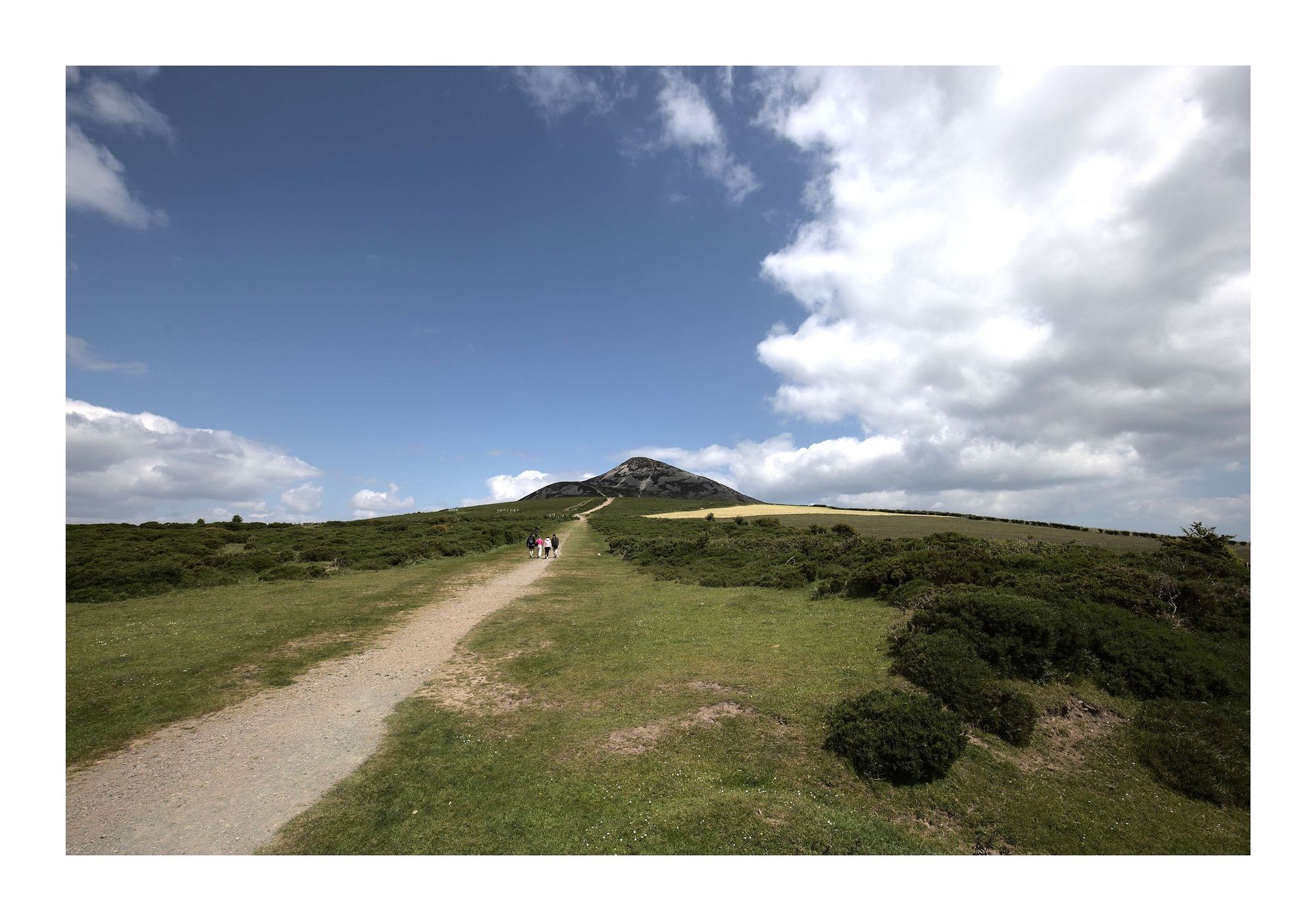 A scenic landscape featuring the Great Sugar Loaf mountain in Ireland. A dirt path winds through the lush green grass, leading toward the rocky peak in the distance. Puffy white clouds fill the sky, creating a striking contrast with the bright blue background. In the middle of the path, a few people can be seen walking, adding a sense of scale to the vast, open landscape.