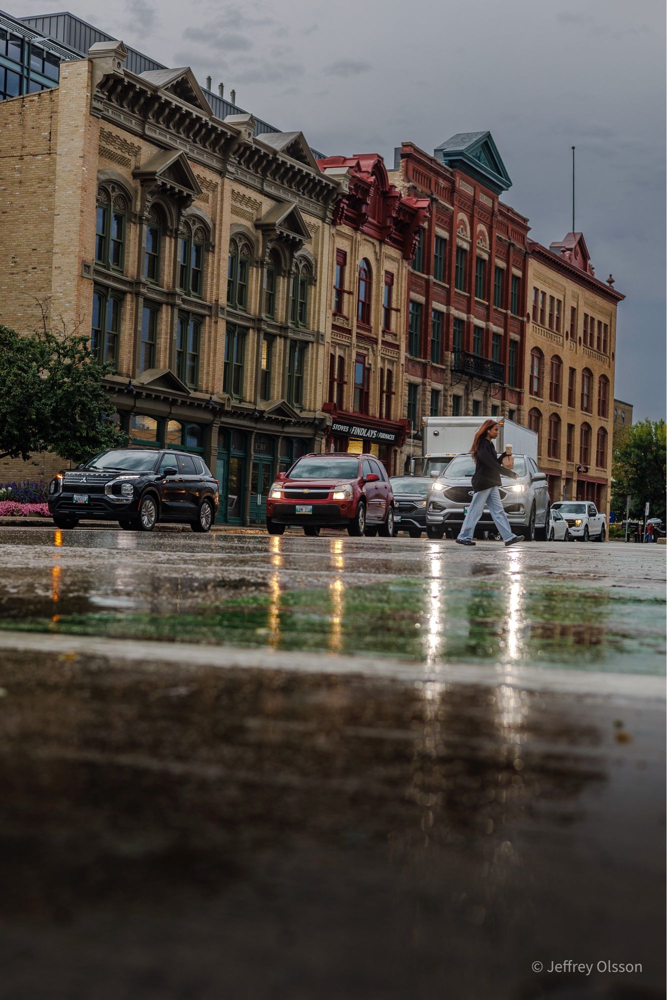 A lady crosses a very wet street while the lights from the cars headlamps reflect off of the road surface.