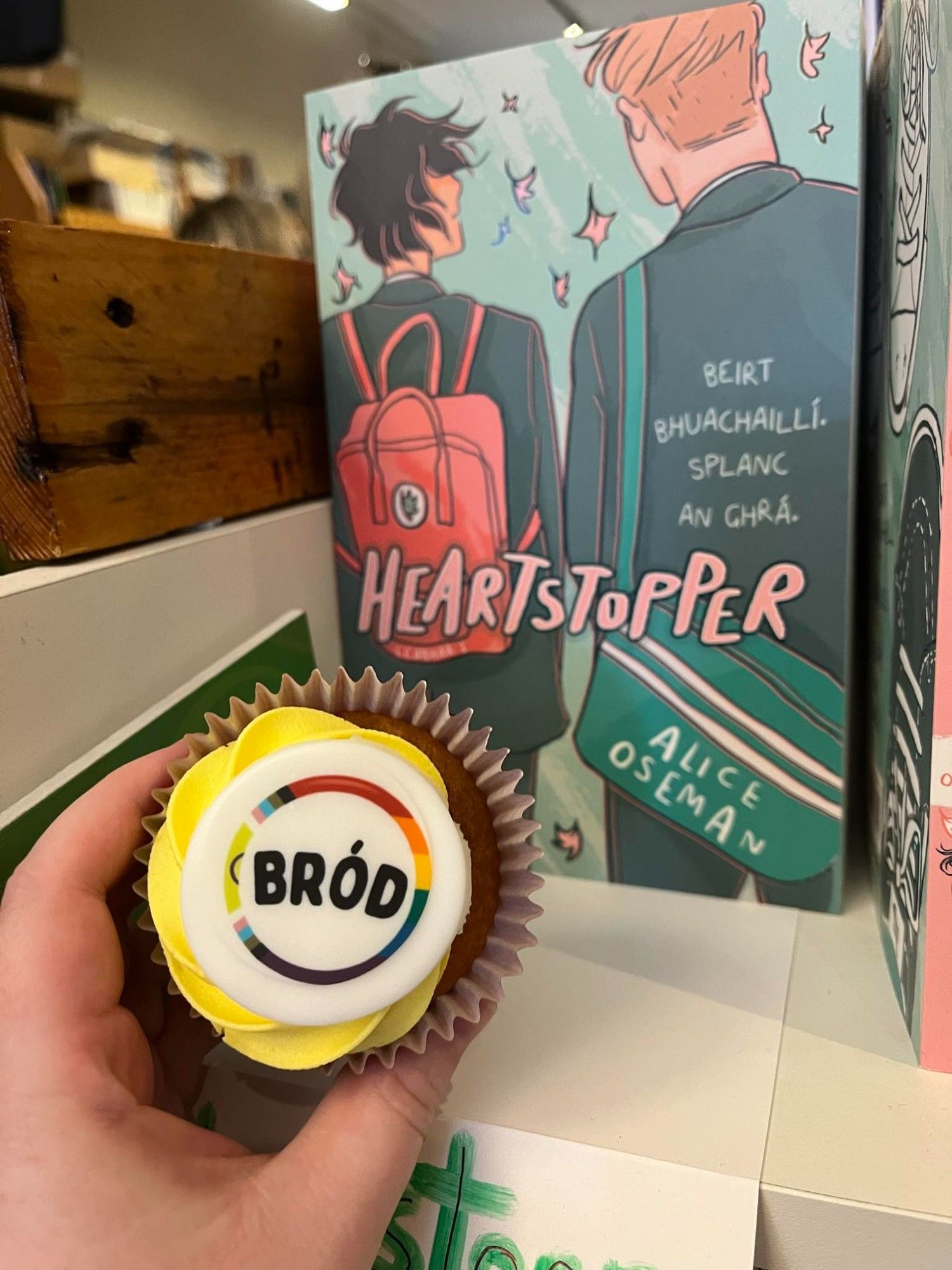 Cístín cupa i lámh ar bun ar clé ar a bhfuil ciorcal bogha báistí a deir Bród, cóip de Heartstopper as Gaeilge sa chúlra / Cupcake with Bród (Irish for Pride) on the top held in a hand in the bottom left corner, in background a copy of Heartstopper in Irish on a shelf.