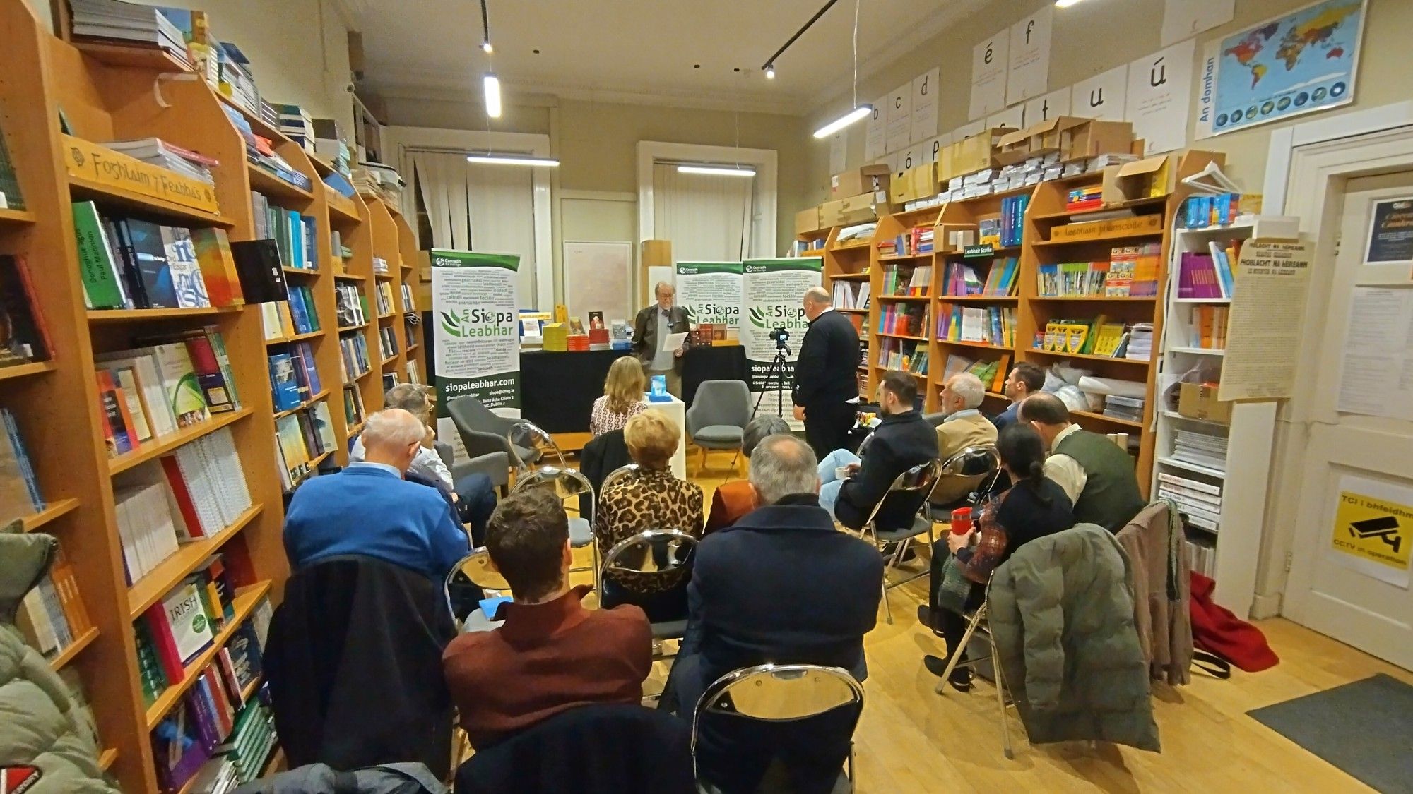 Back end of An Siopa Leabhar [on shuppa lee-y-ower] with crowd of listening people seated to the fore.