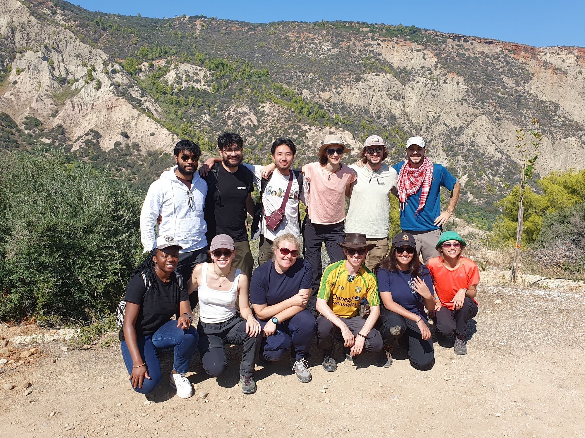 12 smiling doctoral candidates standing in front of a mountain panorama consisting of light coloured rocks, covered by parse green vegetation. The mountain is an uplifted Gilbert delta.