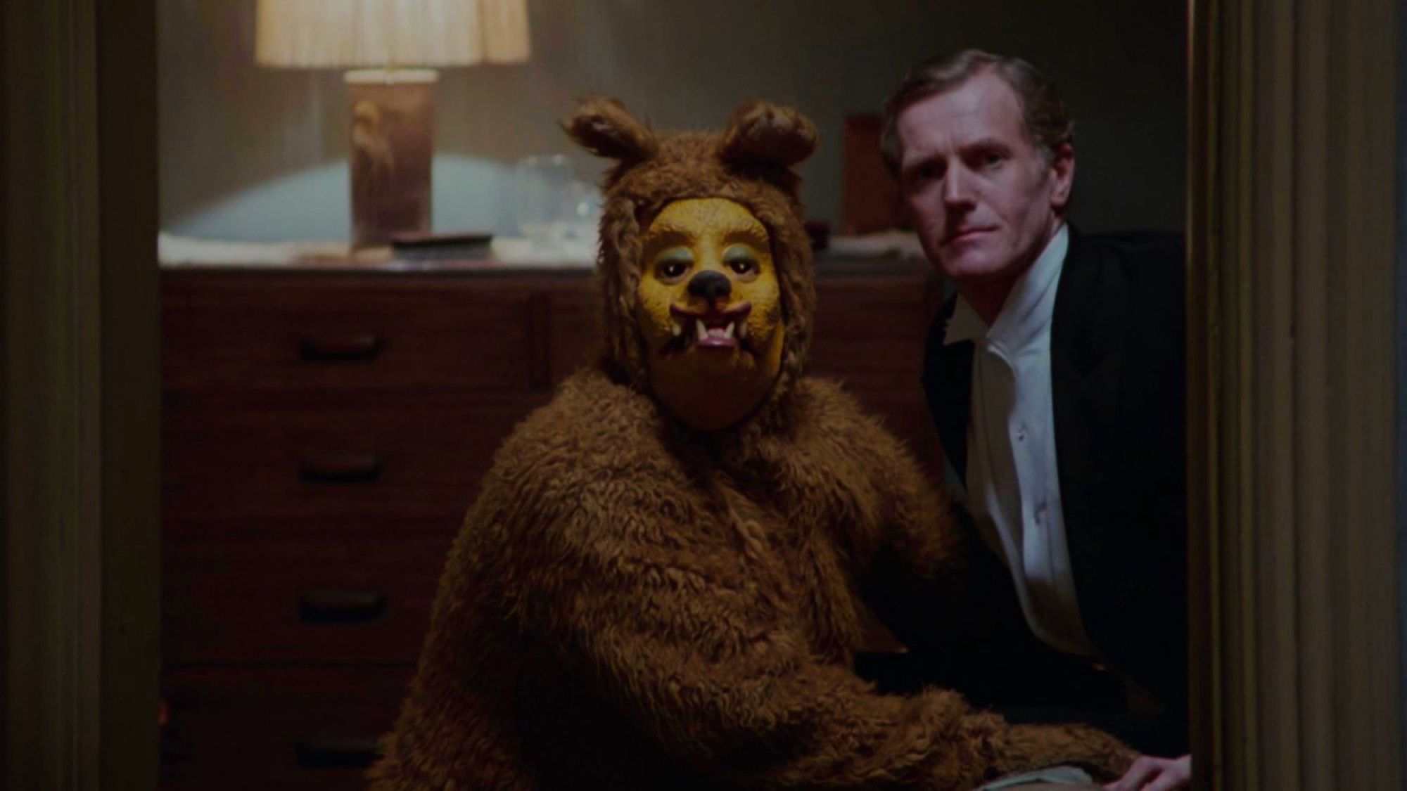 Bear scene from THE SHINING: an older establishment type man sits on a. Bed while someone in a very disturbing bear costume kneels beside him in a too close way that implies something sexual is either happening or just about to happen