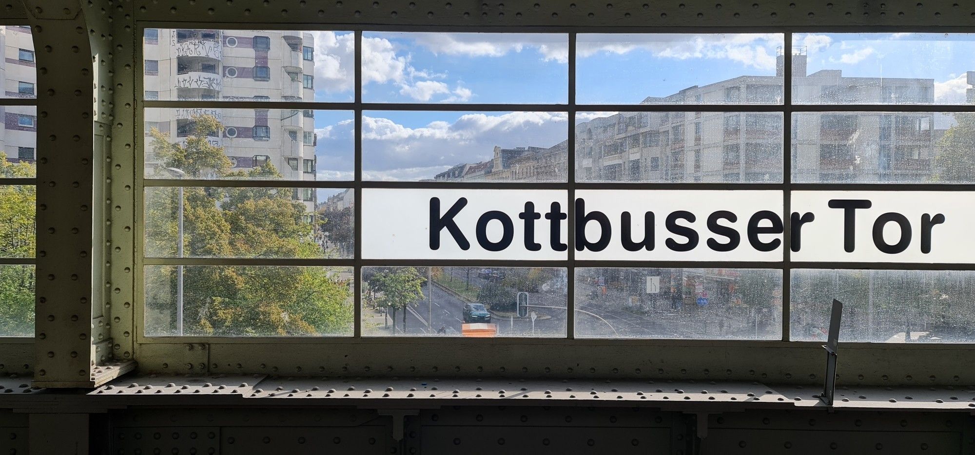 Foto durch die Fenster des U-Bahnhofs Kottbusser Tor. Eine lange Straße und brutalistiche Bauten sind erkennbar.