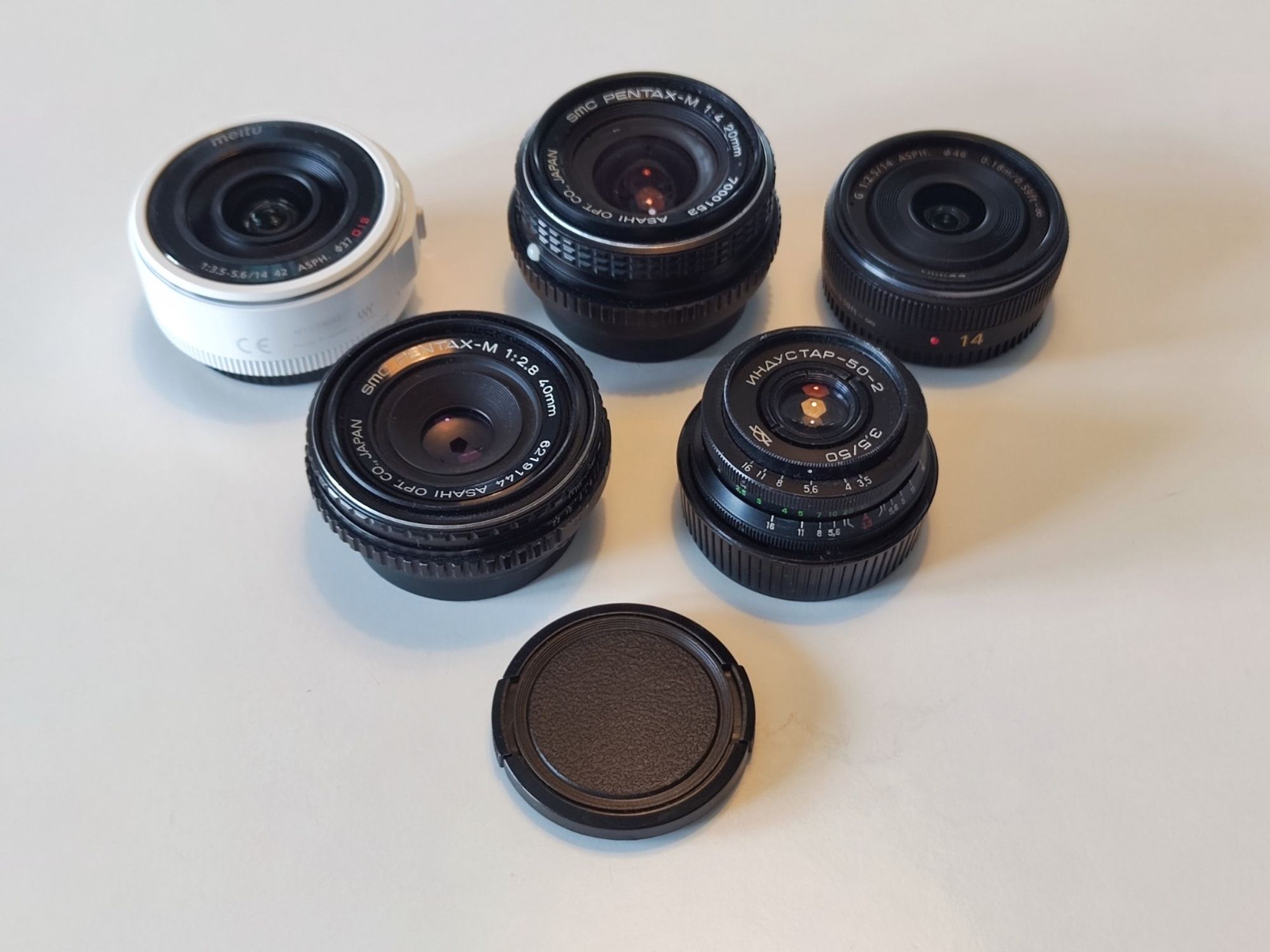 Selection of 5 small lenses: Meitu 14-42mm PZ, smc Pentax-M 20mm, Panasonic G 14mm in the first row; smc Pentax-M 40mm and Industar 50-2 50mm in the second one. 49mm lens cap for comparison.