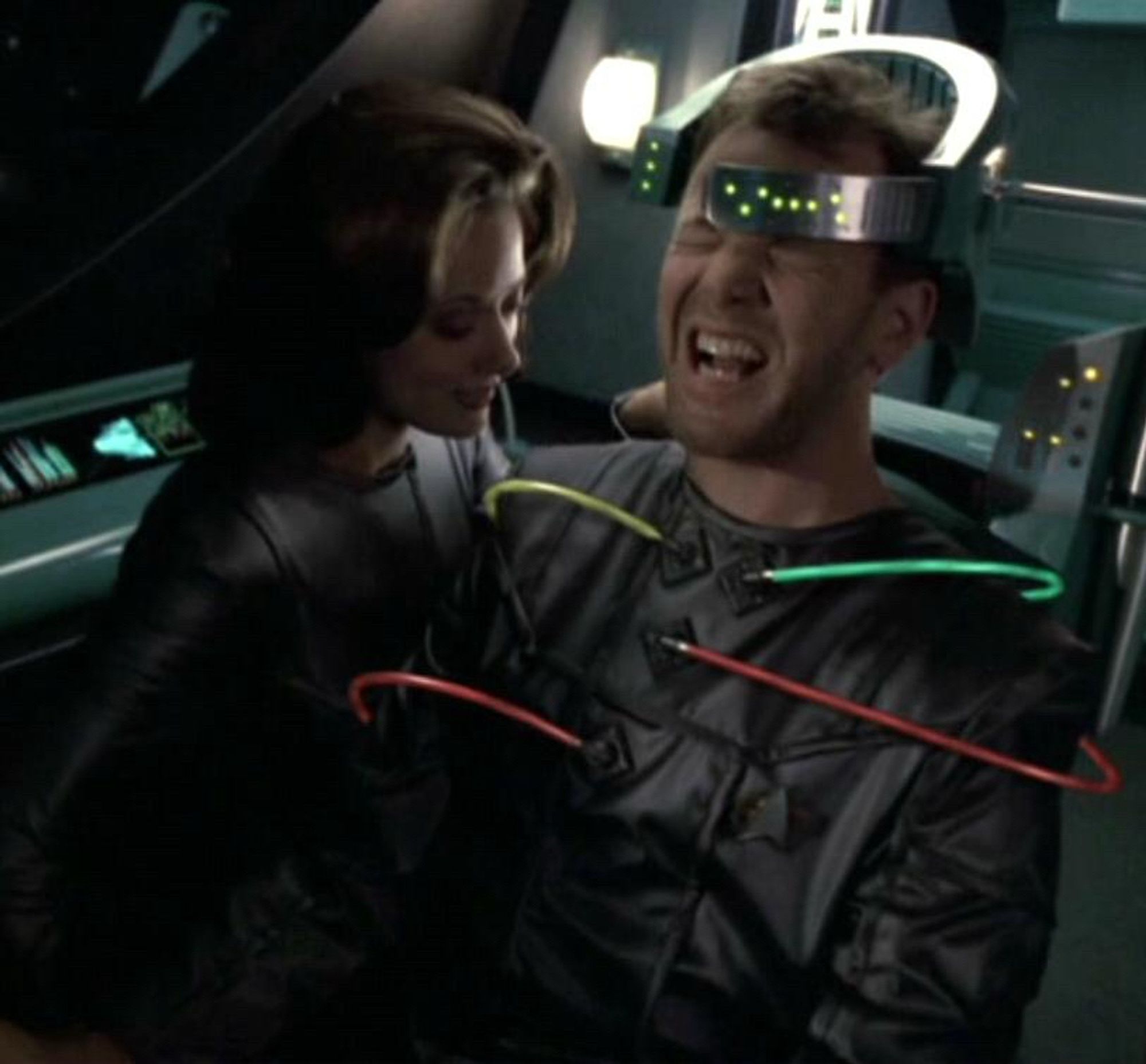 Tom Paris, screaming, is connected to some red, green, and yellow wires with a neural device around the top of his head. Alice, a woman who is also a sentient ship, is looking down on him lustily.