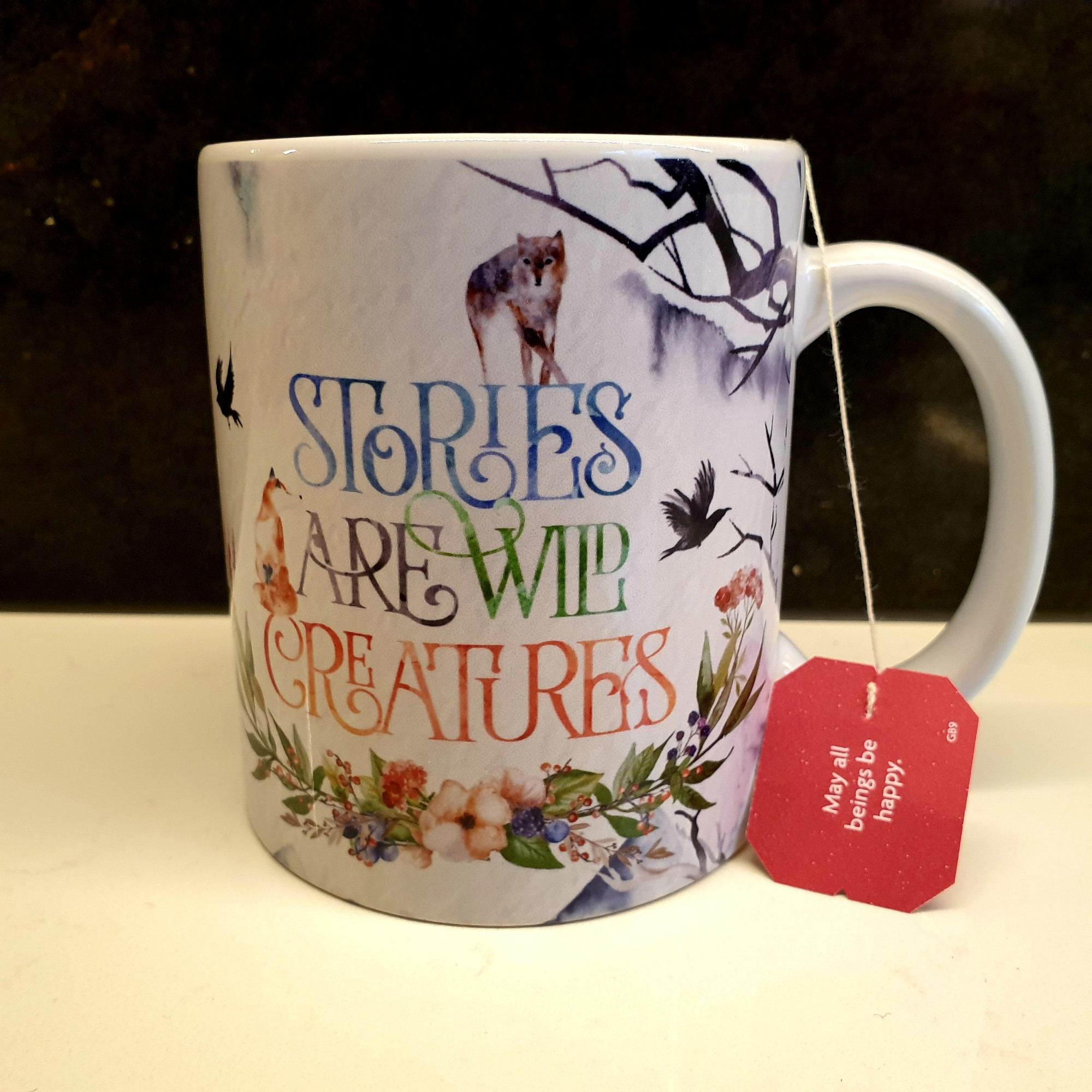 A white mug with the text "Stories are wild creatures" in multiple colours. surrounded by a motif of tree branches, flowers, a wolf, a fox and two crows. There's a tea bag brewing in it, the label on the bag reads "May all beings be happy."