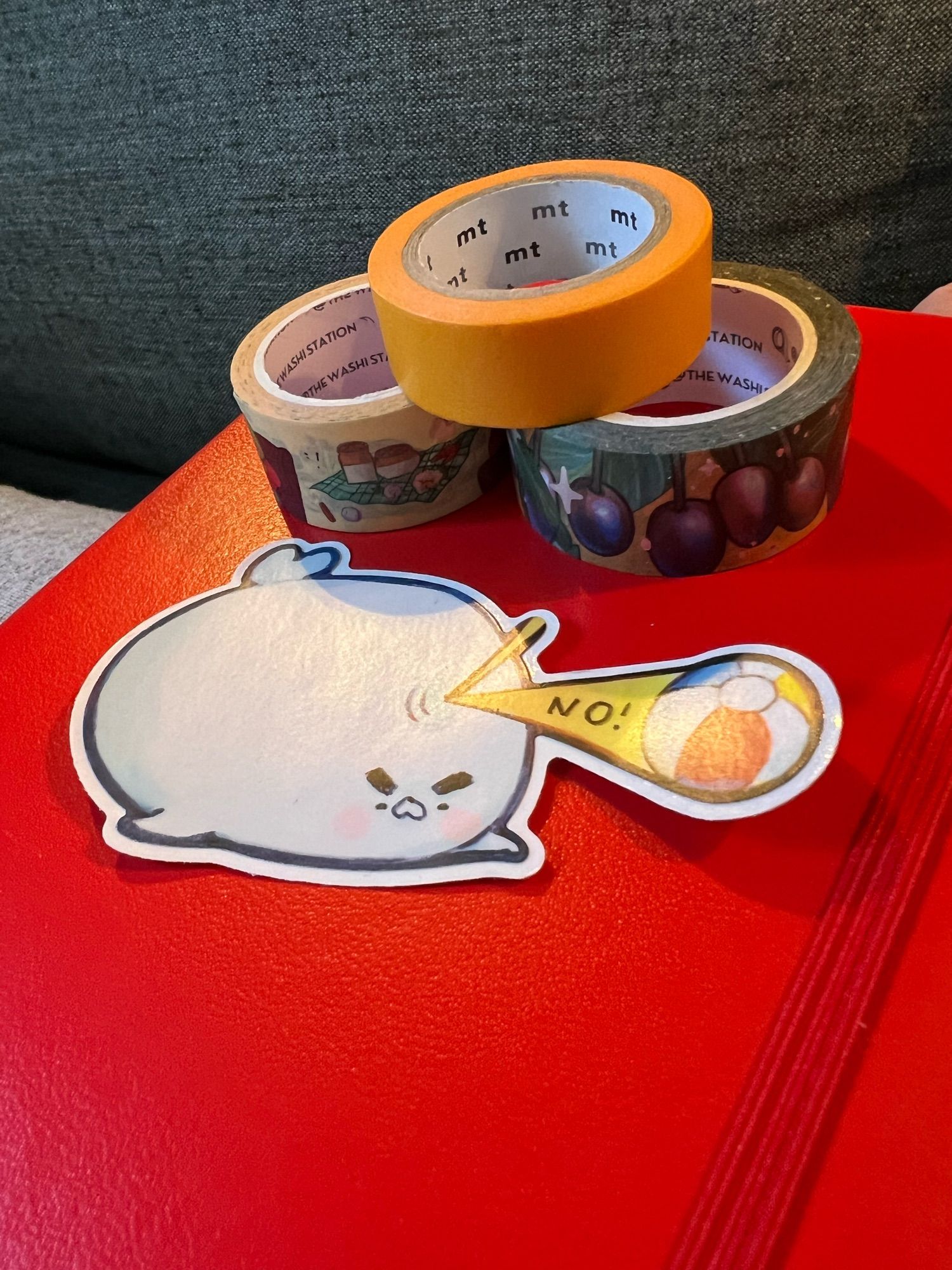 Three rolls of washi tape and a big vinyl sticker on top of a bright red Leuchtturm notebook. The sticker is of a chonky seal bouncing a primary-colored beach ball off it's head and saying "no!" while looking grumpy. The three washi tapes are: a solid golden yellow, a cream background with multi-colored depictions of a cartoon frog living its best domestic life, and a golden background tape with deep purple fruits with green leaves on it.