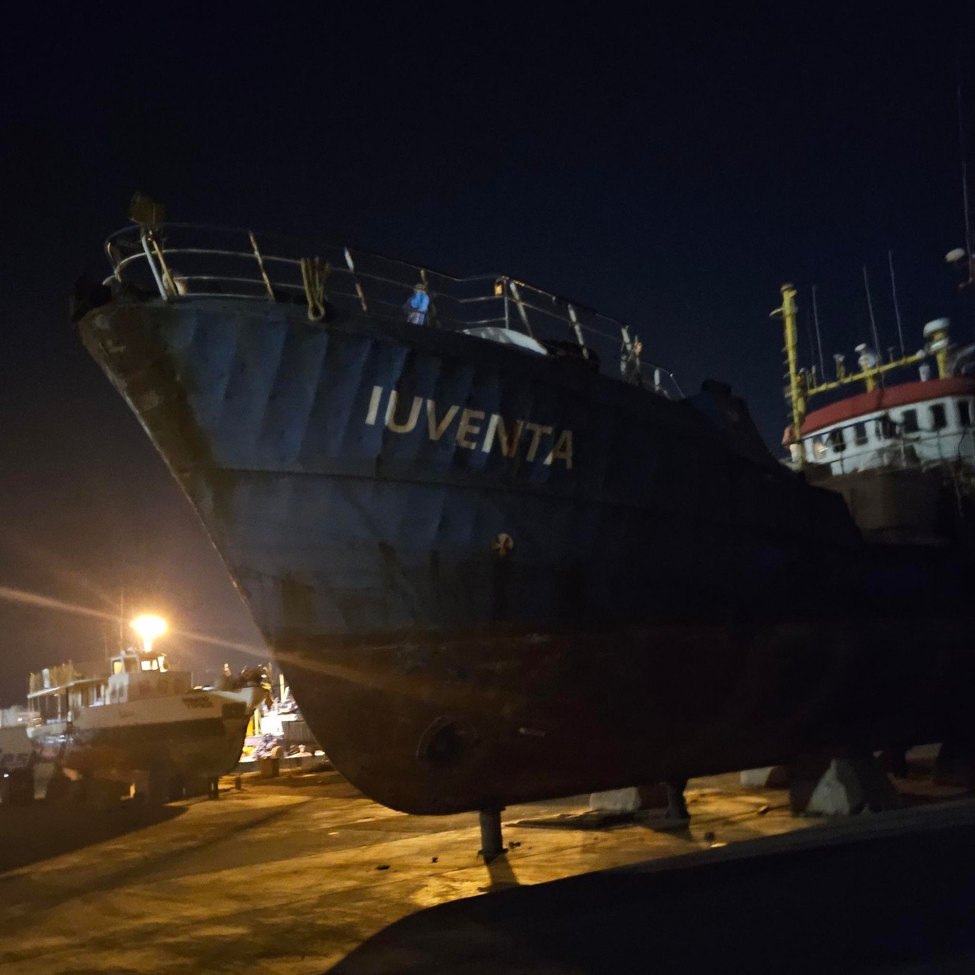 Es ist nacht, links unten im Hintergrund leuchtet ein helles licht. Ein Großteil des bildes ist ein schiff zu sehen, es steht an land. Es ist mit dem bug navh nach Links, im Hintergrund ist die Brücke zu sehen. Am bug steht in großen Buchstaben : iuventa