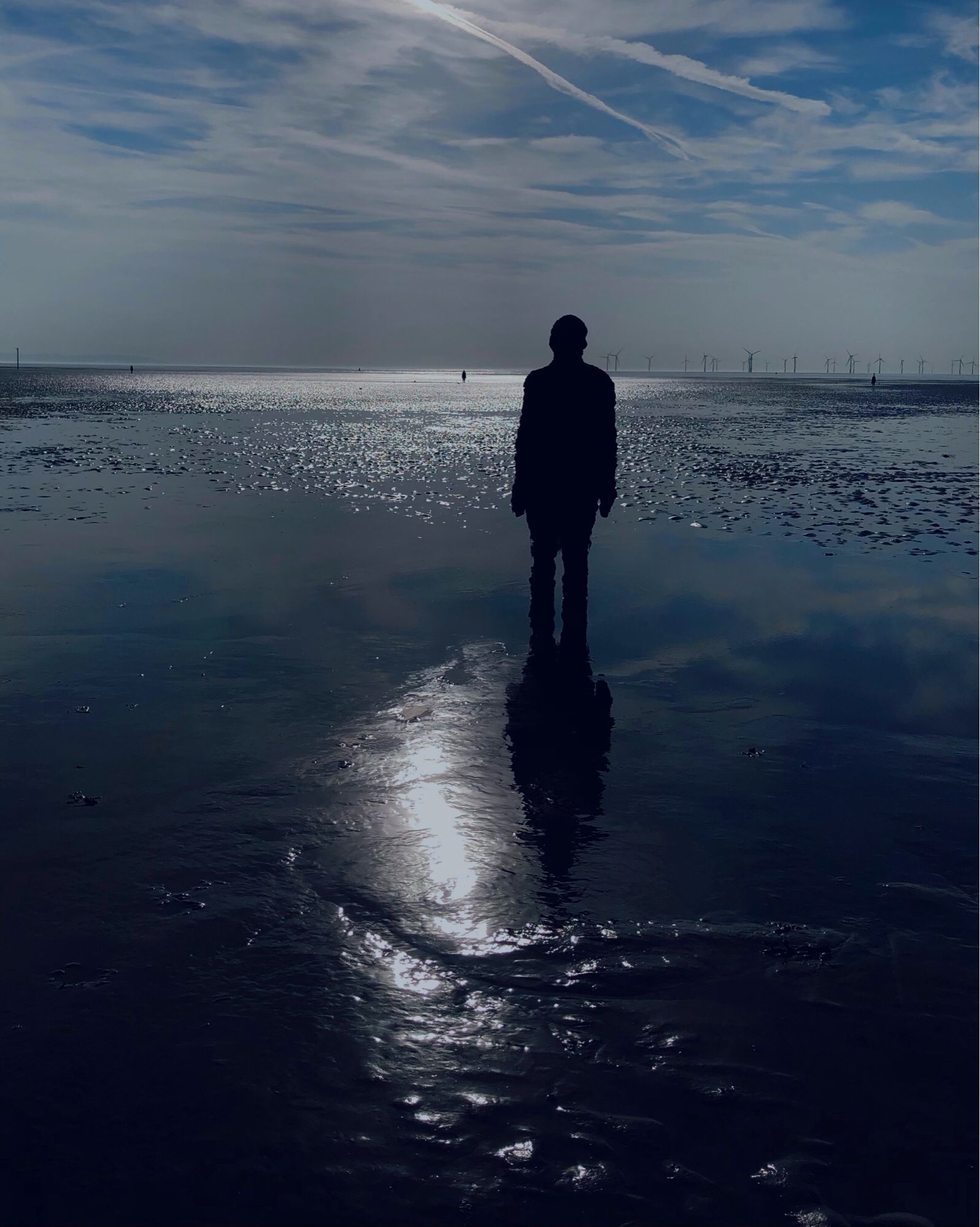 A single bronze Antony Gormley sculpture/figure in the sand as the tide is out with its reflection in the wet sand. The sun catches the sea in the distance giving the appearance of a silvery light pool with a cluster of wind turbines far on the horizon.