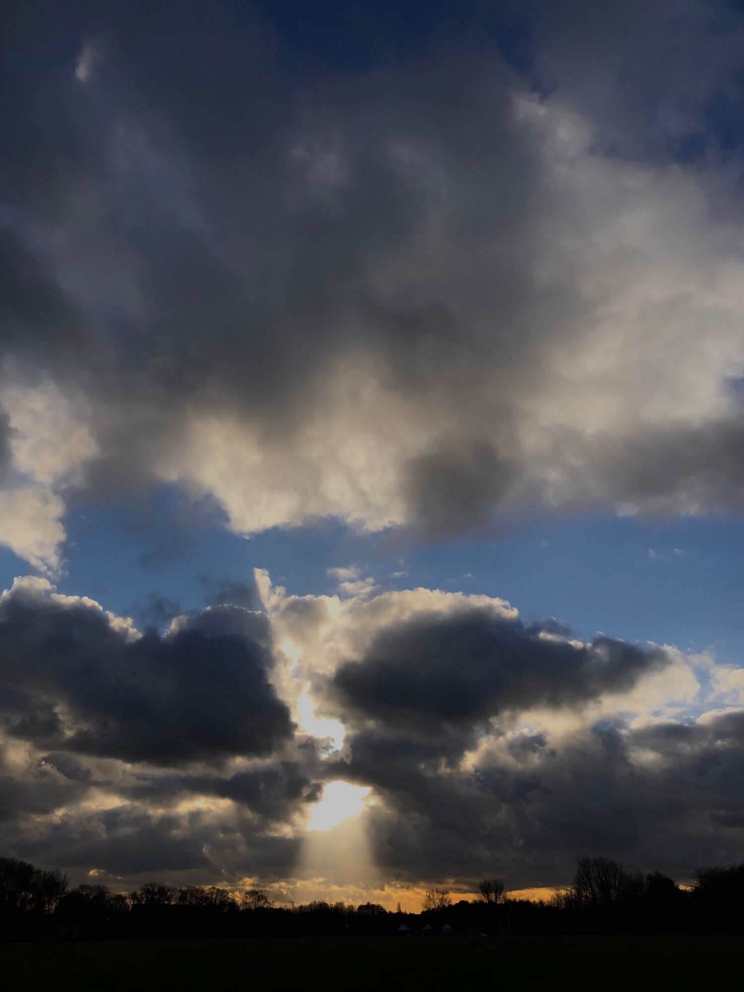 Large grey cumulus clouds before sunset, with a strong shaft of light beaming through a small opening. The horizon is lit with a sliver of orange light & the distant trees black in silhouette.