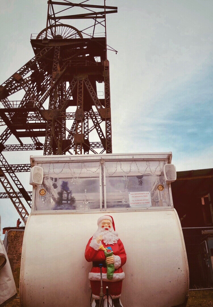 A small caravan with a plastic Father Christmas on the front & small Xmas tree in its window.
Behind looming large the rusted structure of a Pit Head & wheel.