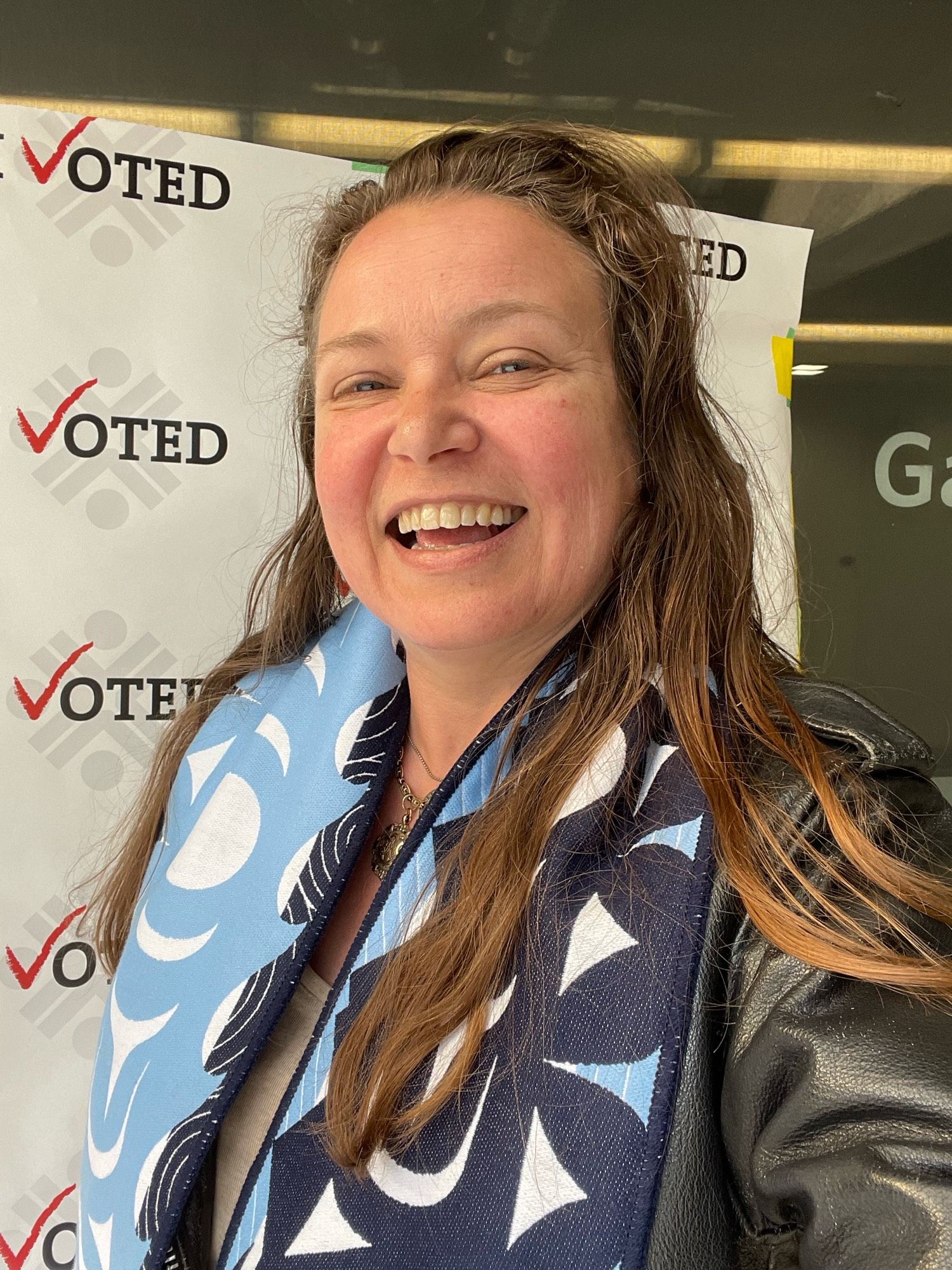 Smiling lady in Vancouver Whitecaps Salish design scarf posing in front of ‘I Voted’ poster